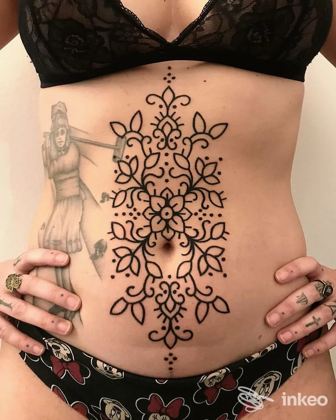 Stomaco di una persona apparentemente femminile con tatuaggio ornato nero, incisione centrale verticale e motivi floreali, accompagnato da altri tattoo sui lati, sfondo neutro.