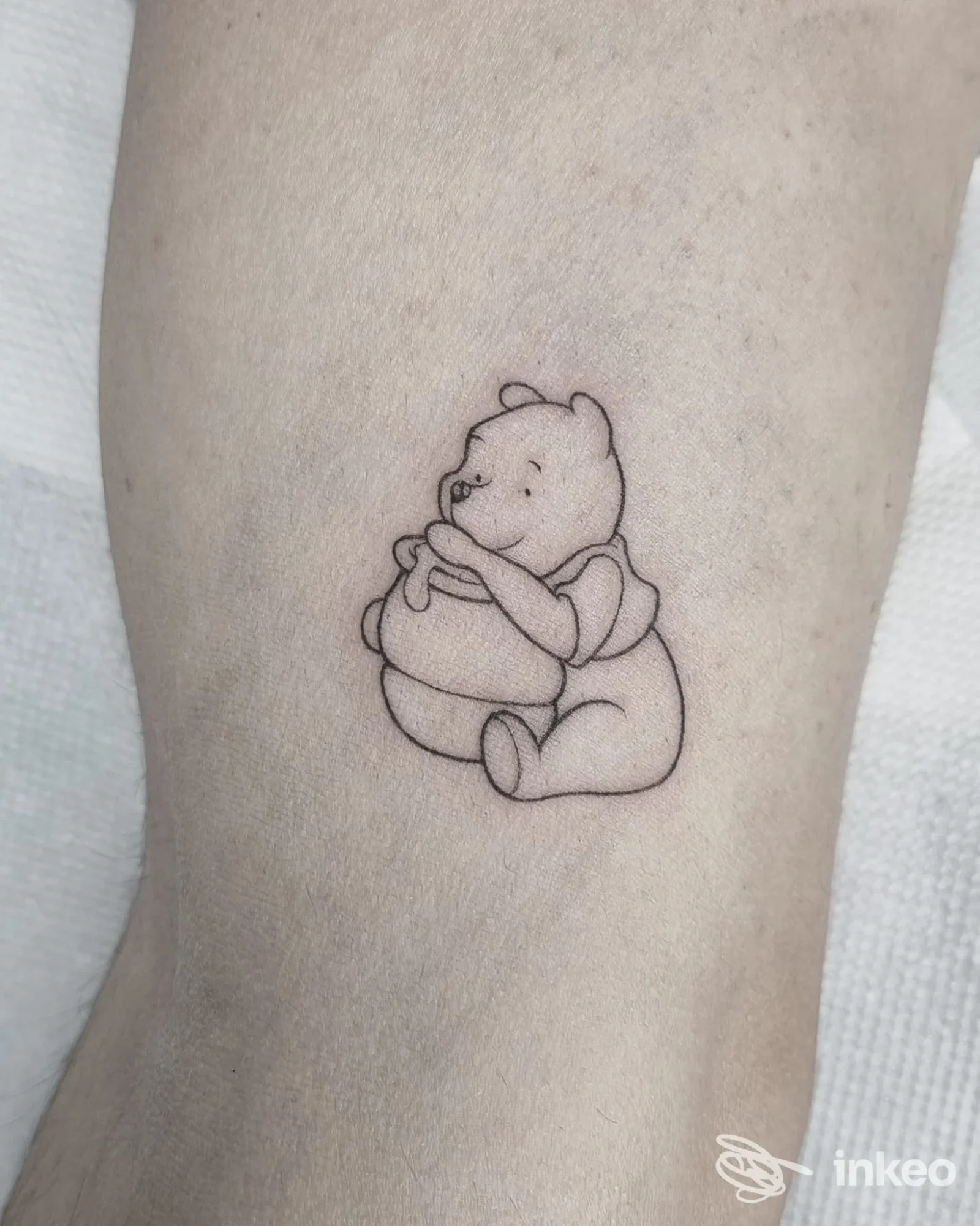 Tatuaggio su pelle raffigurante Winnie l'Ourson seduto, disegnato in linee nere, visibile sull'avambraccio.
