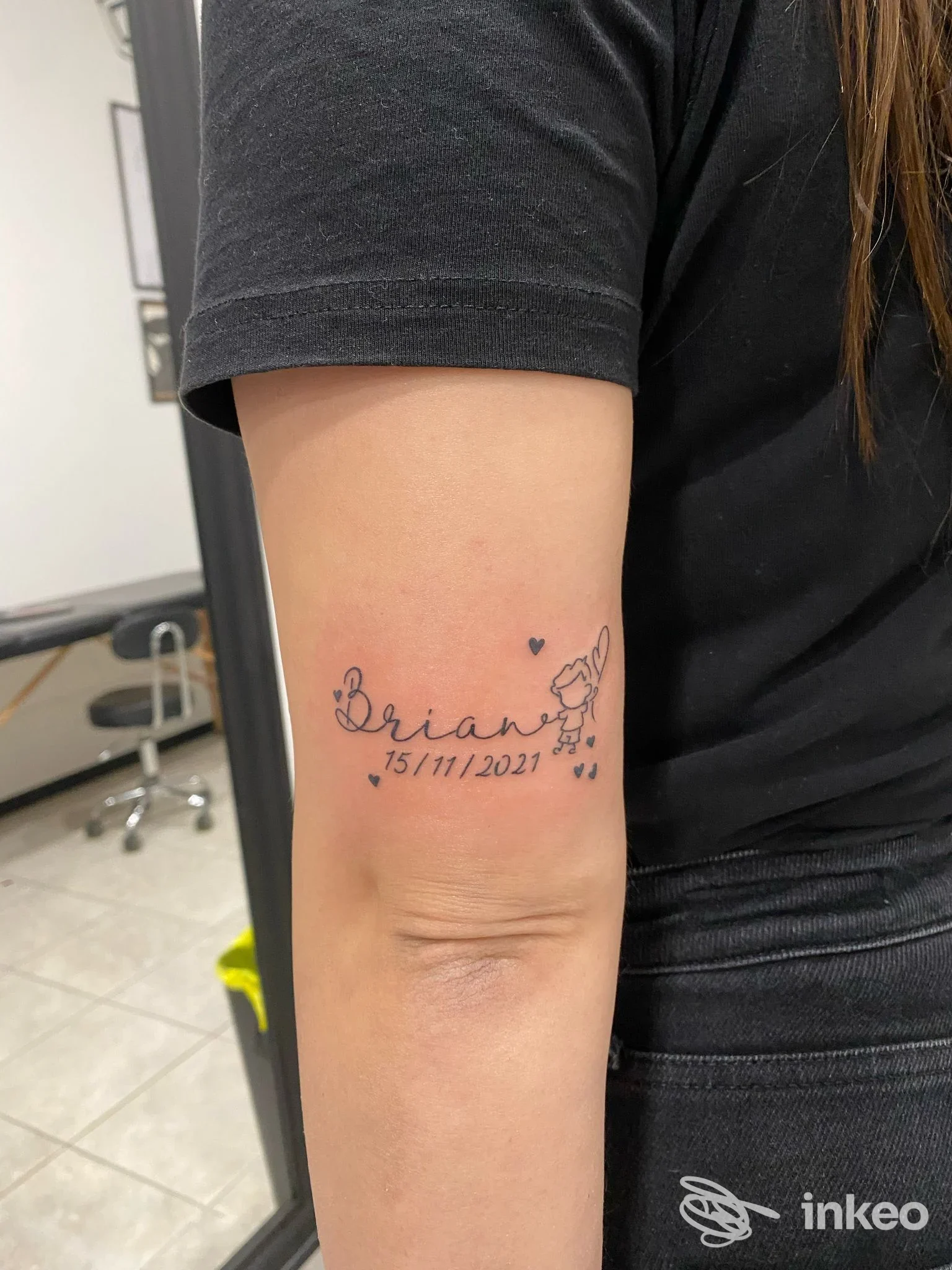 Foto dell'avambraccio di una persona con un tatuaggio: nome scritto, cuori e piccoli fiori, data 15/11/2021. Nessun volto visibile, contesto studio, semplice e nitido.