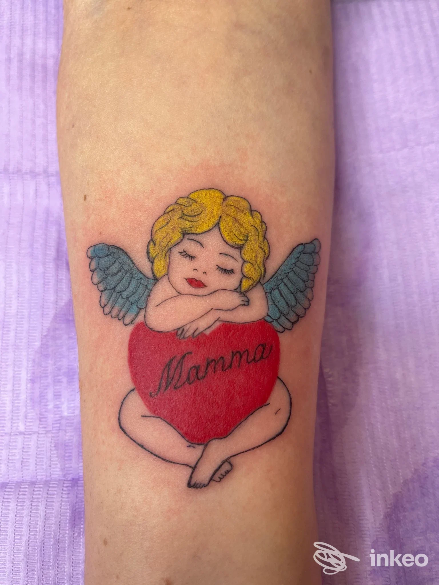Tatuaggio su avambraccio di un angelo con ali azzurre che tiene un cuore rosso con la parola Mamma, senza visione di volto reale.