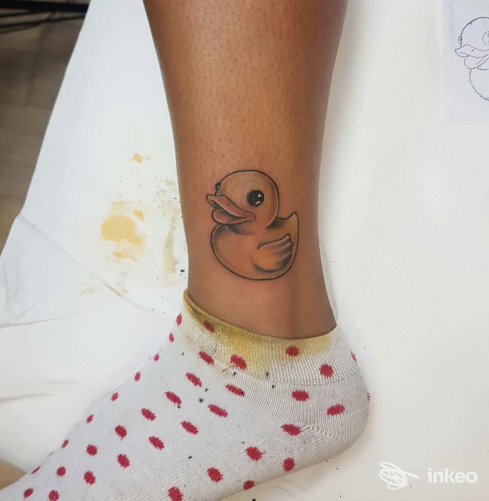 Tatuaggio semplice di una paperella arancione sulla caviglia di una persona, con contorni definiti e riempimento uniforme.
