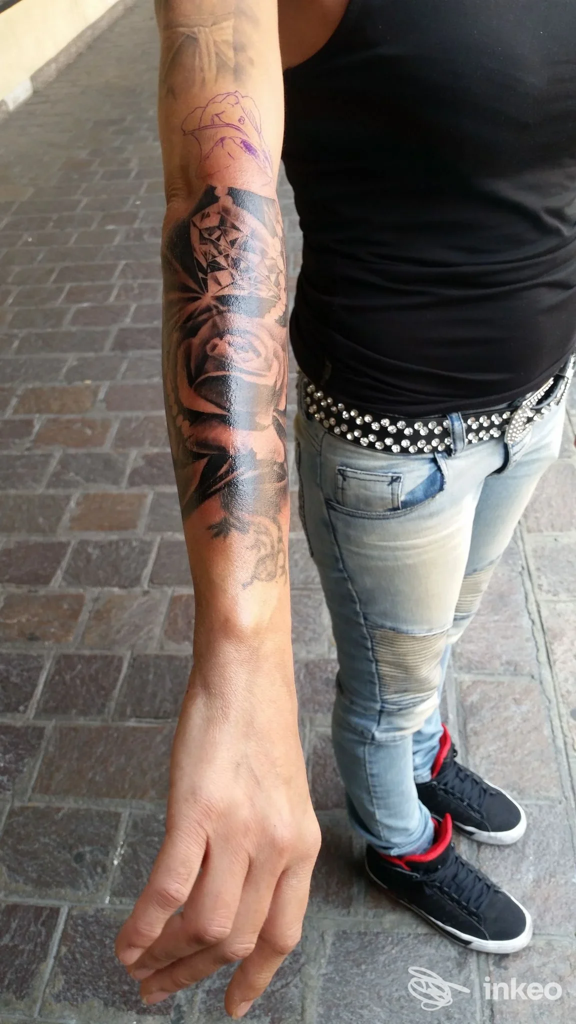 Esteso avambraccio di una persona con tatuaggio nero scuro lungo la pelle, indossa maglietta nera e jeans, sfondo urbano.