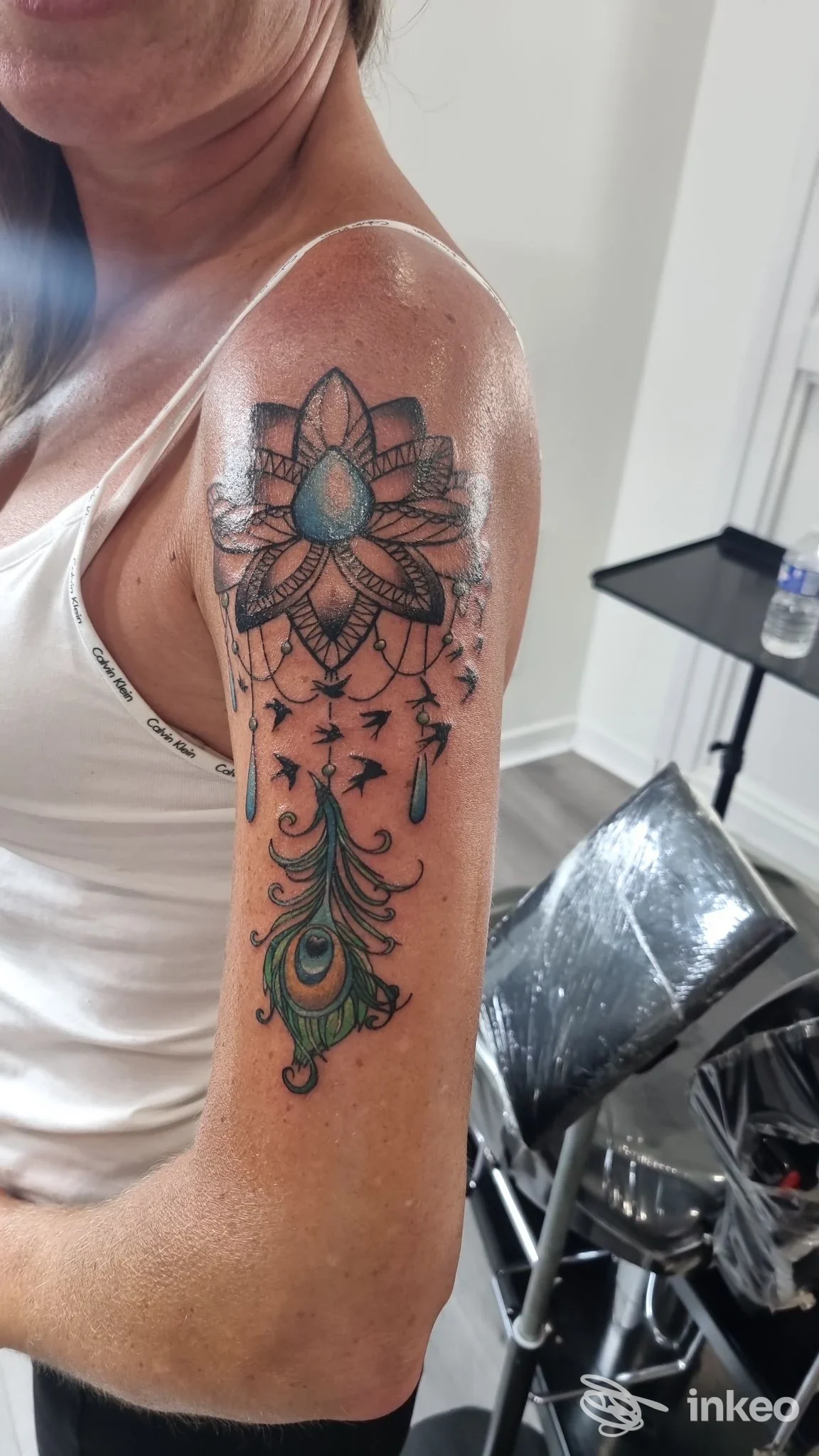 Tattoo sul braccio sinistro con lotus e motivi mandala, decorazioni verdi a pennacchio lungo la parte superiore del braccio, visibile in studio.