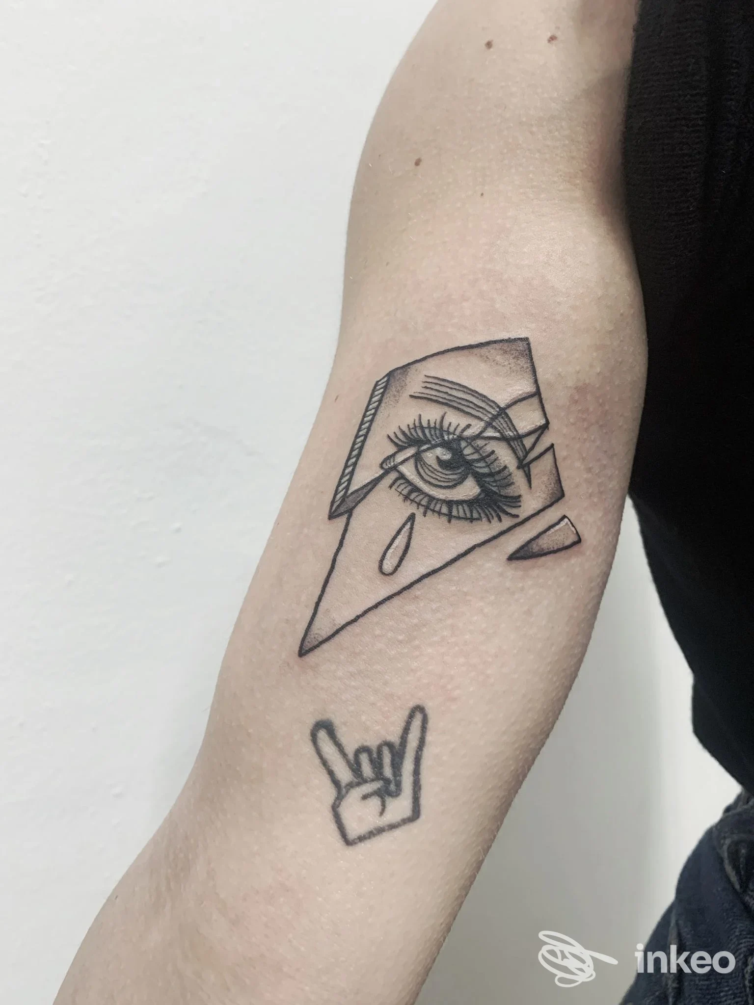 Tattoo sul braccio: disegno geometrico con occhio e lacrima, accompagnato dal piccolo gesto rock on. Sulla pelle si intuisce una foto scattata in studio. (P'ink Tattoo Studio)