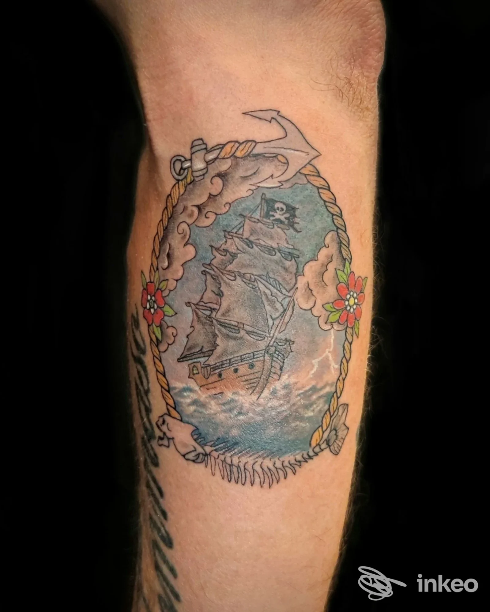 Tatuaggio su avambraccio raffigurante una nave a vela entro cornice ovale di corde, con nuvole e fiori sui lati; realizzato sulla pelle.