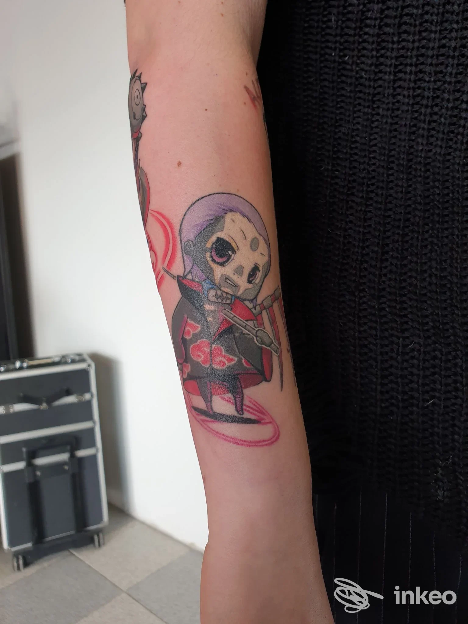 Tatuaggio sul braccio raffigurante una piccola figura scheletrica in abiti scuri con dettagli rossi e contorni neri.