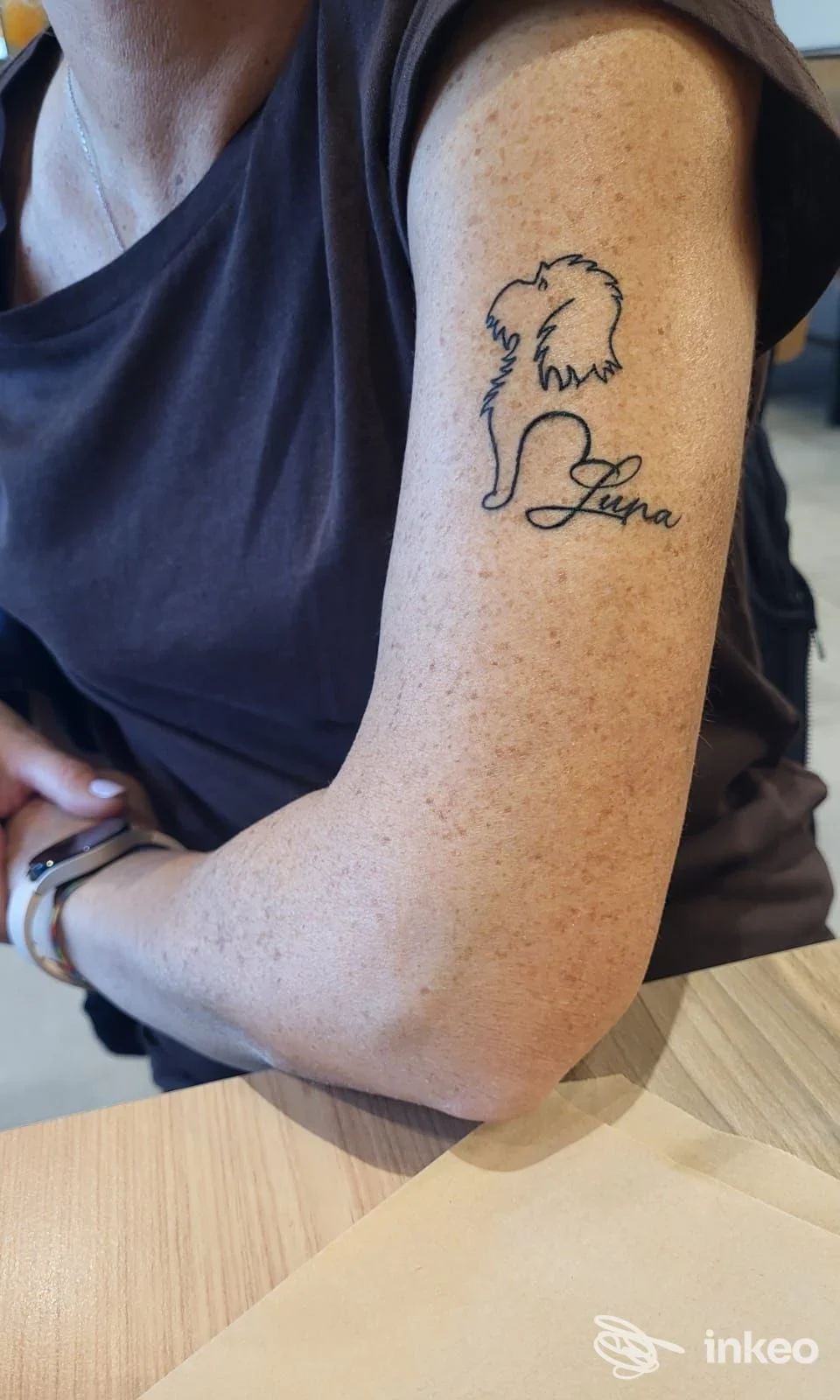 Braccio di una persona con un tatuaggio a linea nera di un cane stilizzato e una scritta decorativa; pelle leggermente punteggiata, abbigliamento manica corta, sfondo neutro.