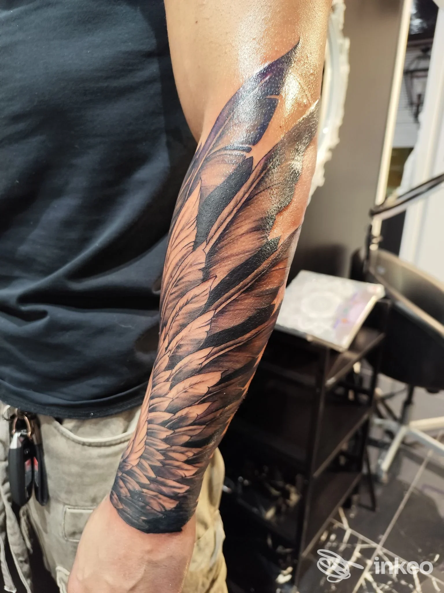 Tattoo a manica sull'avambraccio di una persona, motivo di piume scure. Nessun volto visibile e sfondo dello studio.