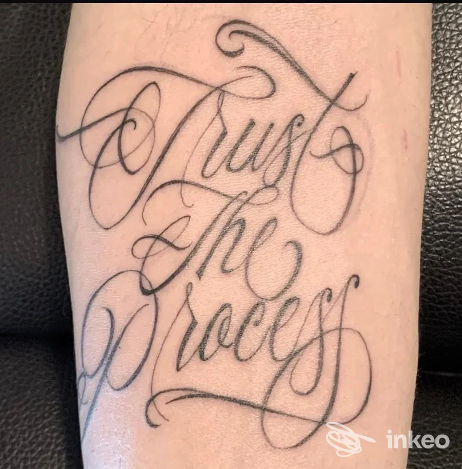 Tatuaggio su avambraccio: scritte ornate in stile corsivo che leggono «Trust The Process», realizzato direttamente sulla pelle.