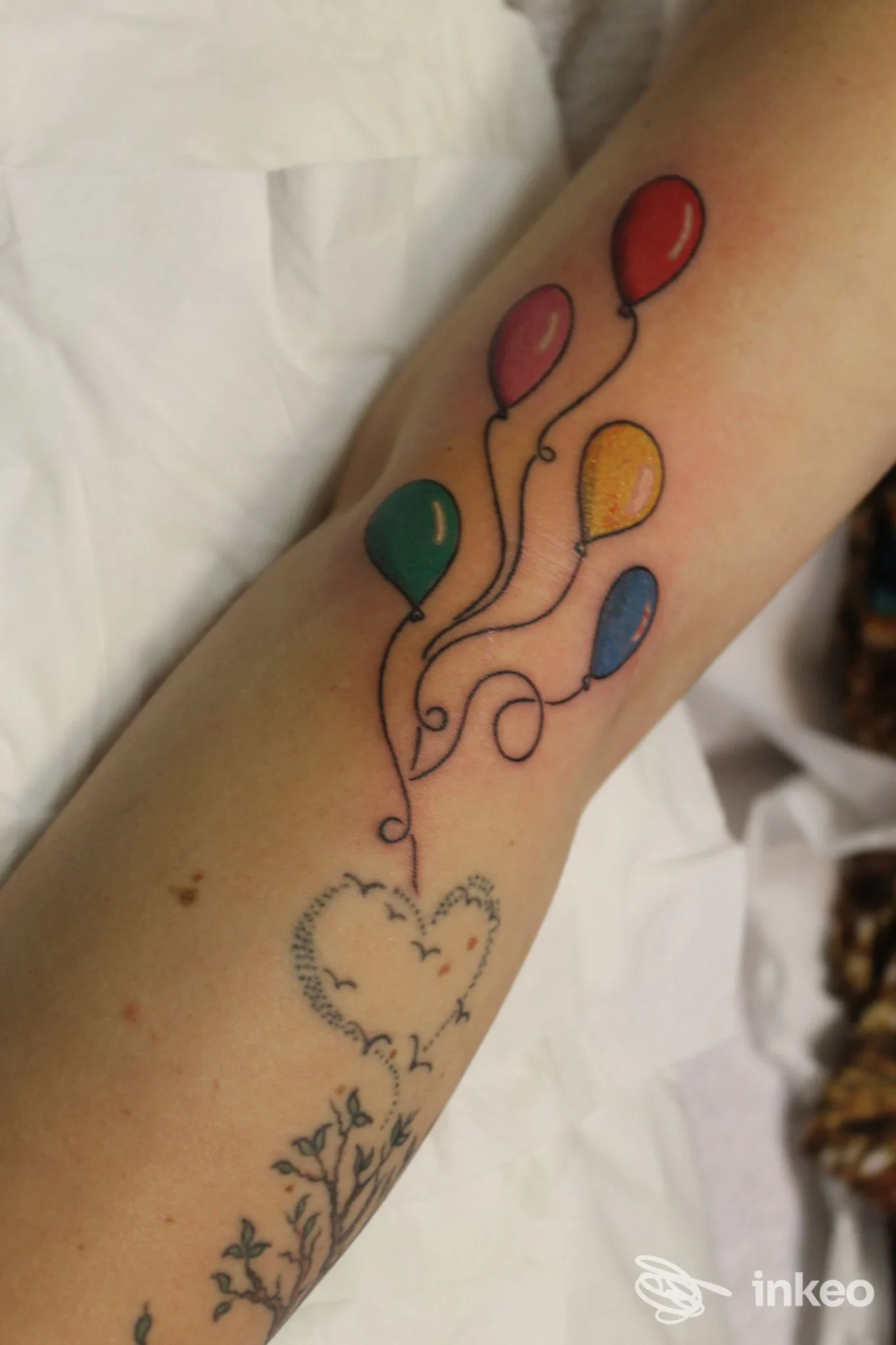 Avambraccio con tatuaggio colorato di palloncini stilizzati su rami che escono da un cuore tratteggiato; i palloncini sono rossi, arancione, verde, blu e viola, con contorni neri ben definiti.