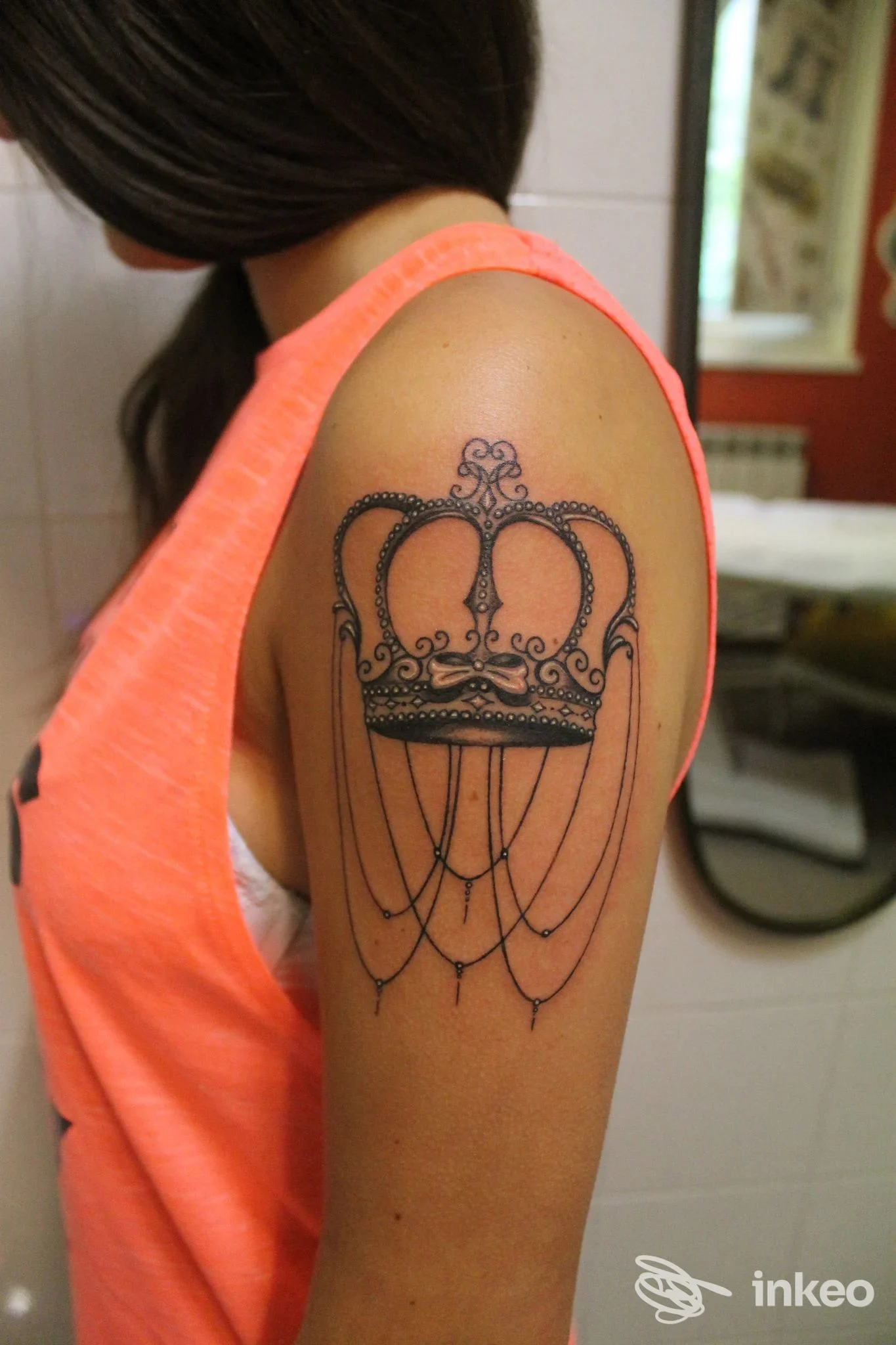 Tatuaggio su braccio raffigurante una corona con pendenti a cascata, visibile chiaramente su una spalla di una persona in studio, senza volto visibile.