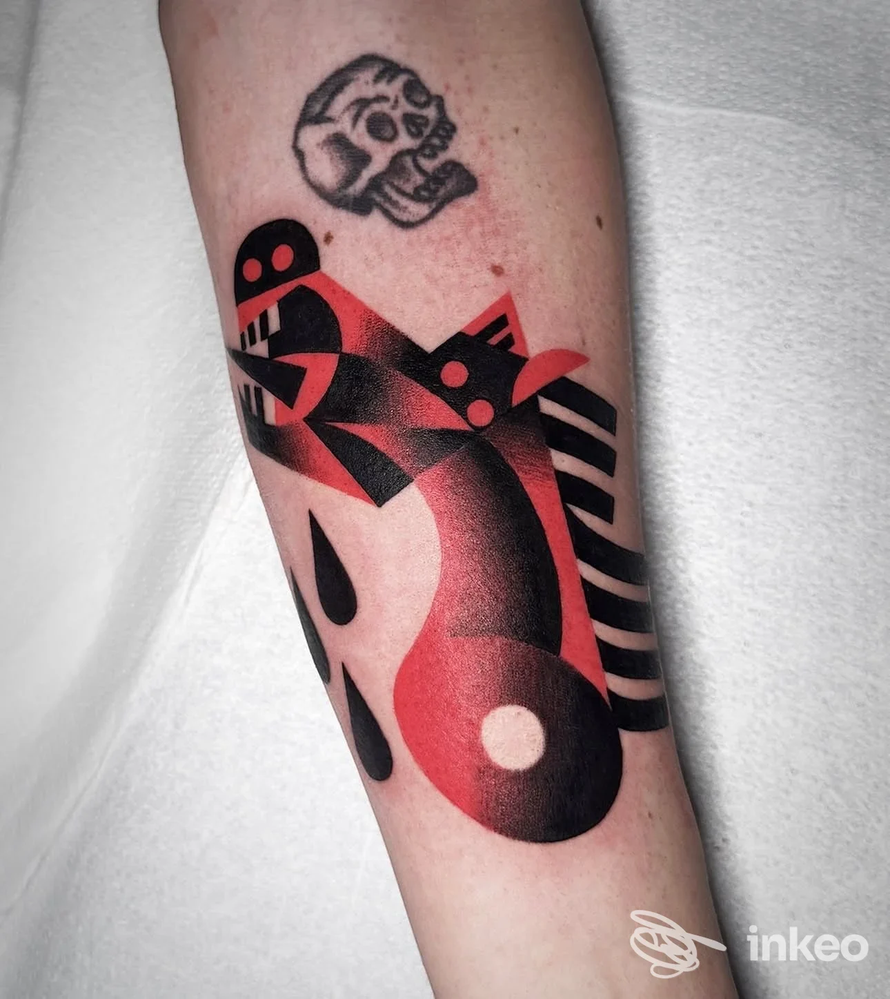 Tattoo finito su avambraccio: figura geometrica rossa e nera con dettagli a linee, accompagnata da un piccolo teschio in alto, su pelle reale.