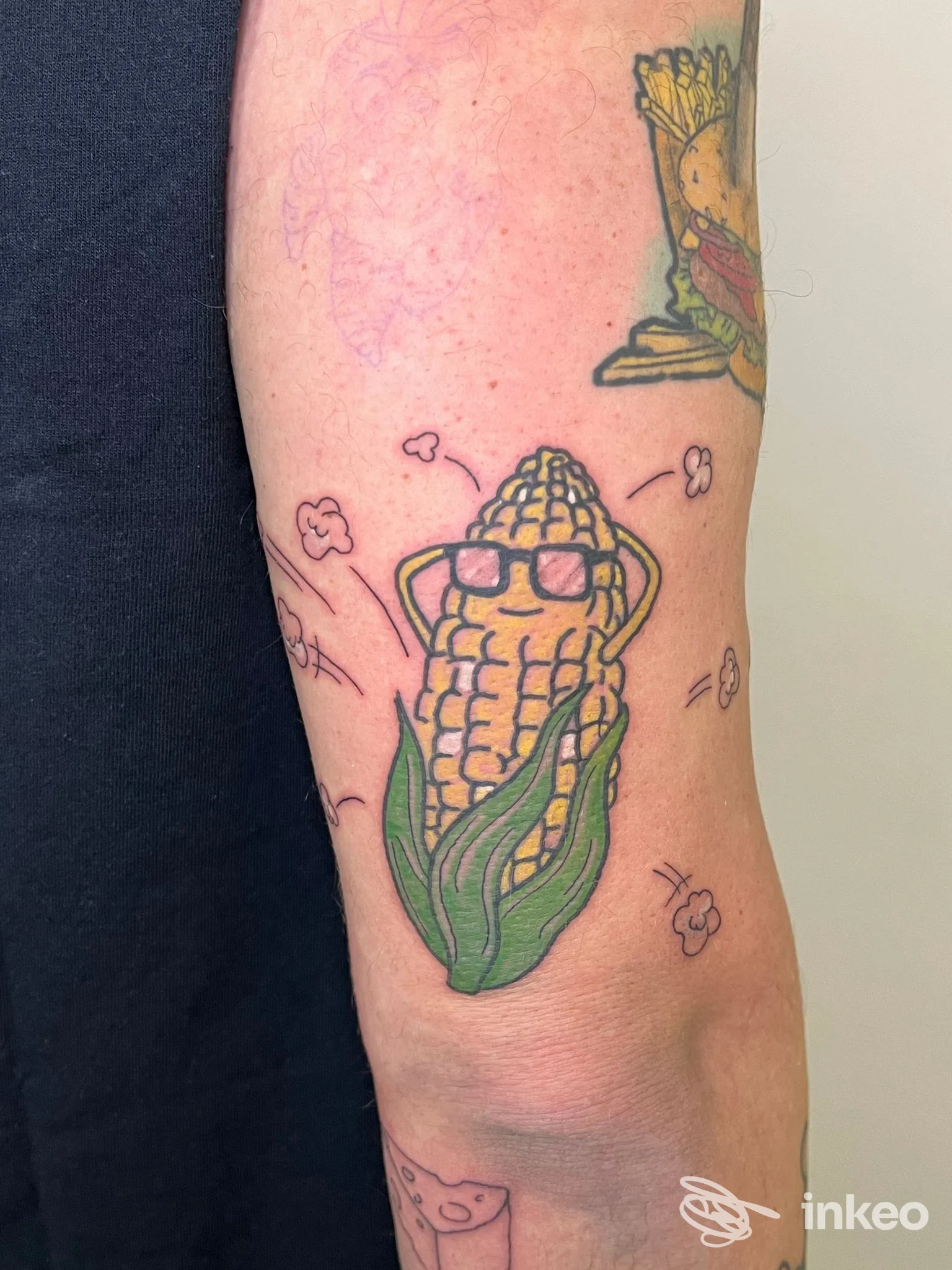 Tatuaggio su avambraccio di un mais stilizzato con occhiali da sole, colorato e ben delineato.