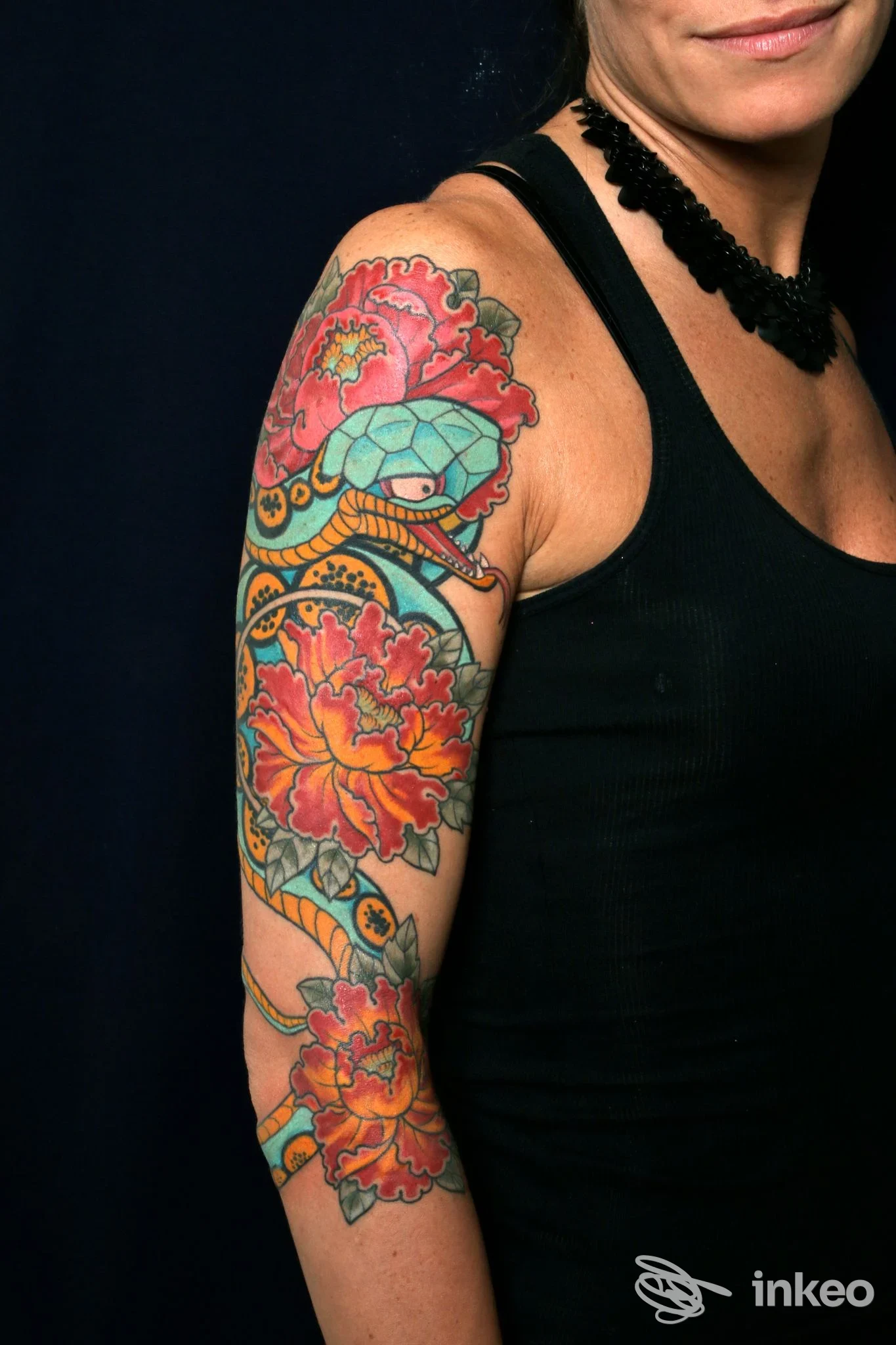 Braccio di una persona con tatuaggio colorato di fiori e serpente, visibile parzialmente dal busto in su, sfondo scuro, con canotta nera.