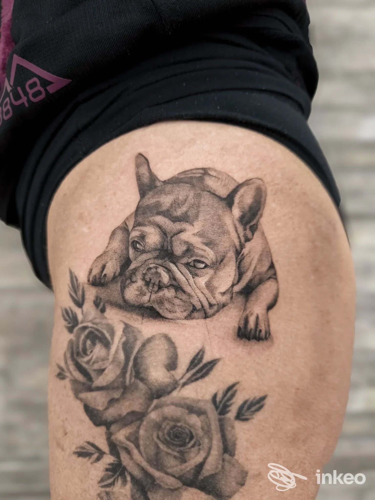 Tattoo visibile sulla coscia di una persona: un bulldog stilizzato in tonalità grigie, accompagnato da rose, dettagli di shading puliti su pelle.