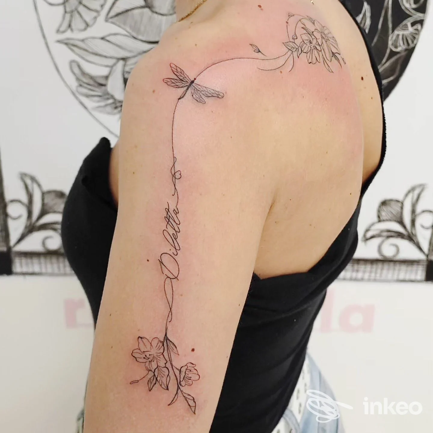 Tattoo a linea sull'avambraccio con fiori e una scritta elegante lungo il tratto verticale, visibile su pelle chiara senza volto mostrato.