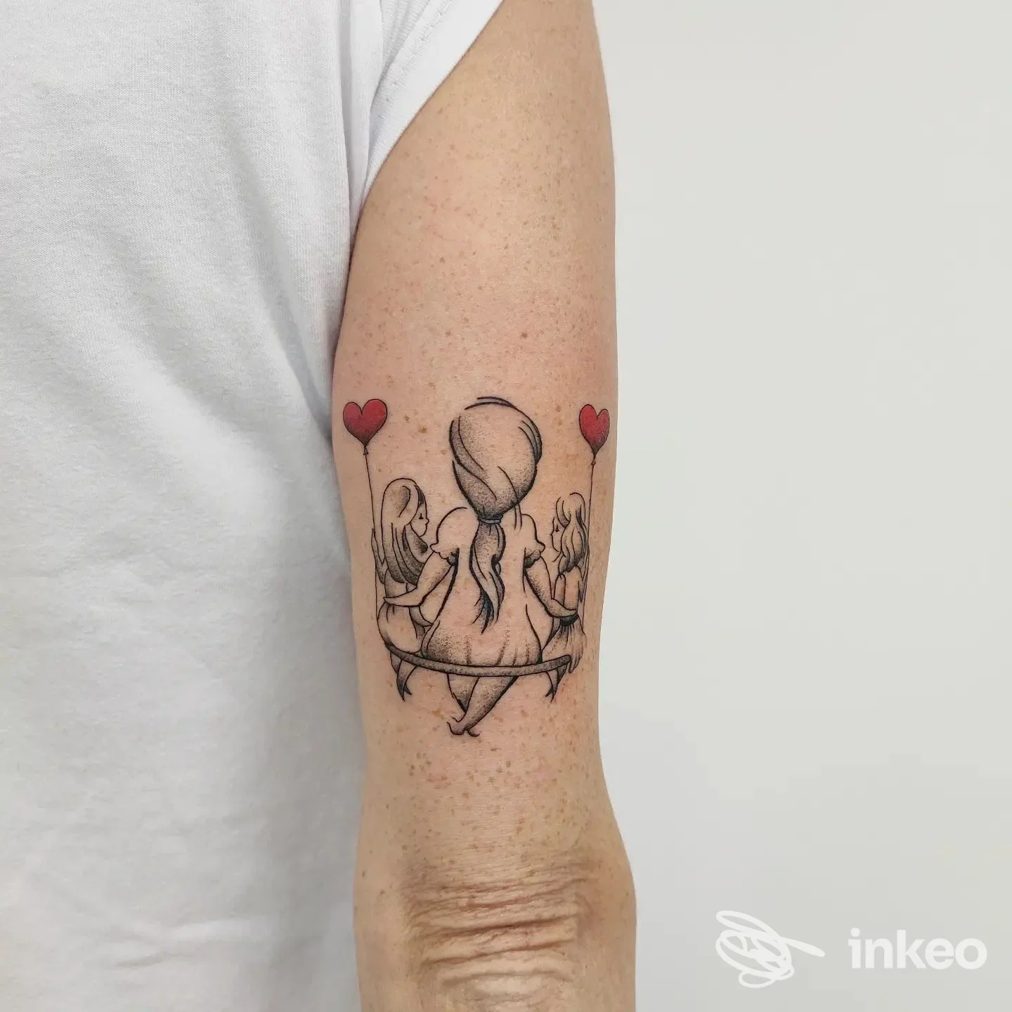 Tatuaggio sul braccio raffigurante una figura centrale seduta su una panchina, accompagnata da due figure ai lati, e tre cuori rossi sospesi come palloncini.