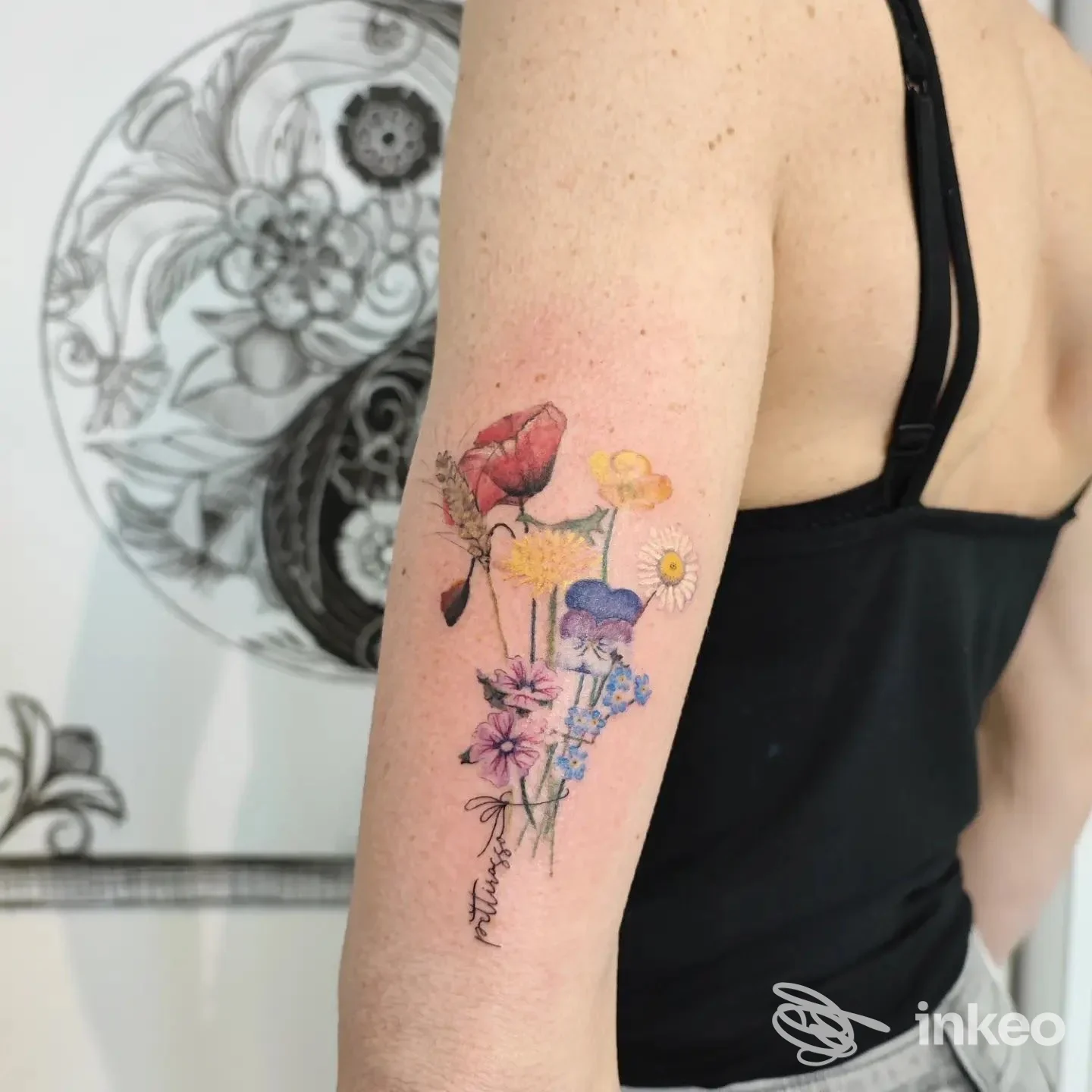 Tattoo floreale colorato visibile sul braccio di una persona, con fiori in tonalità rosse, gialle, blu e viola. Nessun volto visibile.
