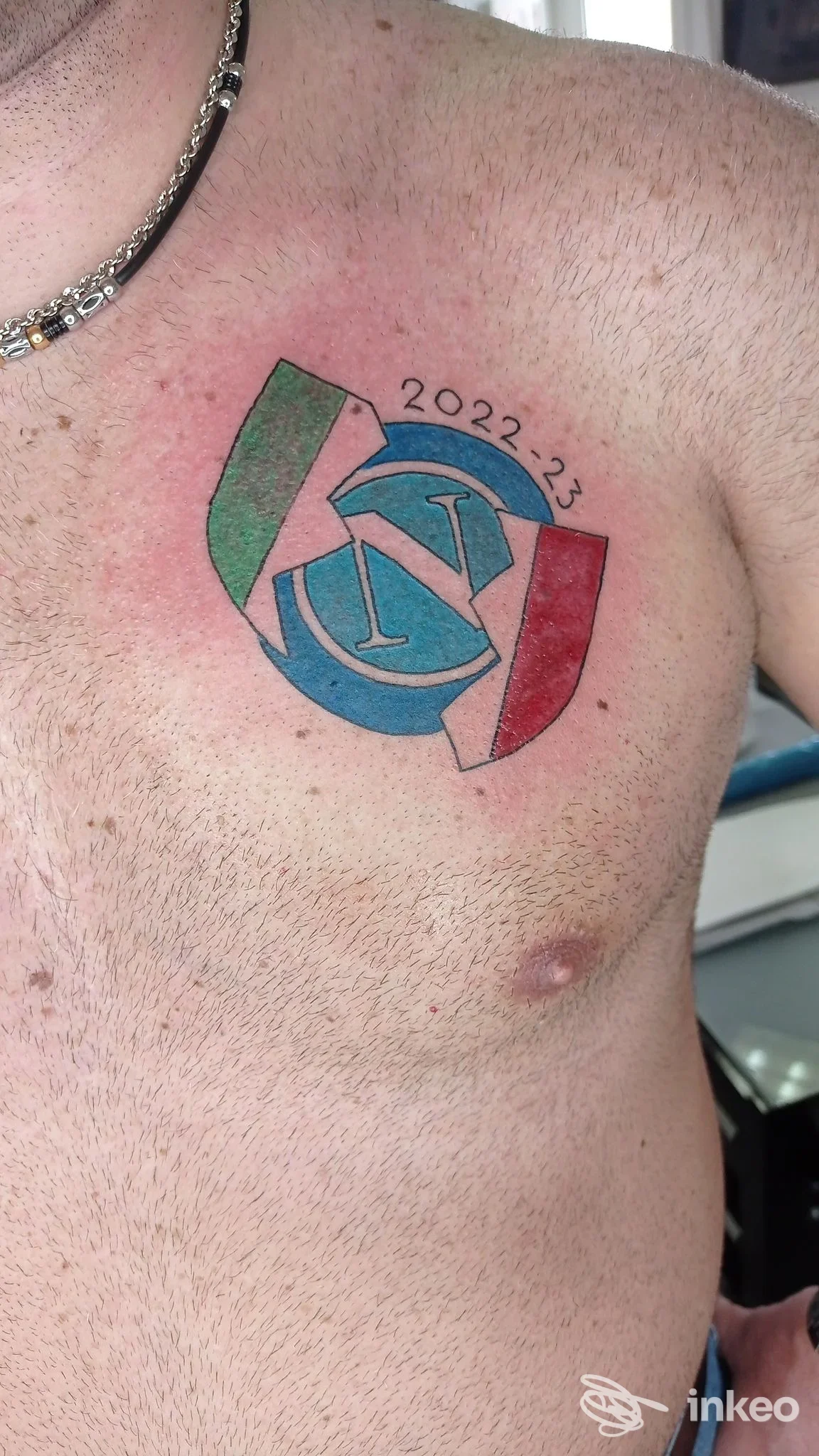 Tattoo a colori sul petto sinistro, circolare con bande verde, bianca e rossa e testo 2022-23; pelle visibile senza watermark o overlay. Luke's Tattoo Studio, luogo non specificato.