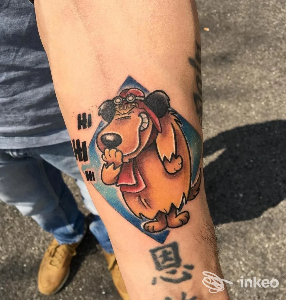 Tatuaggio a colori su avambraccio raffigurante un cane in stile cartoon con cappellino, realizzato direttamente sulla pelle, visibile lateralmente.