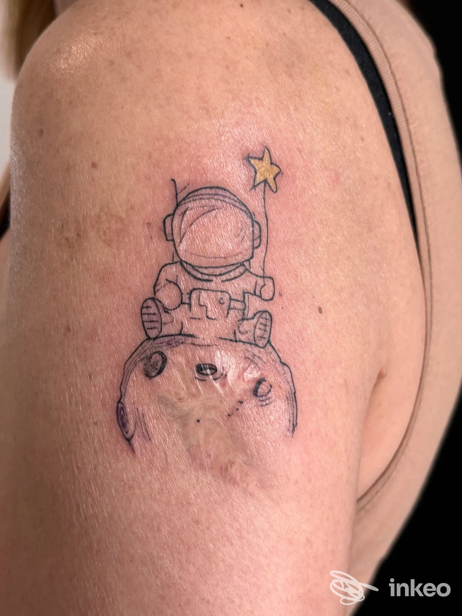 Tattoo visibile sul braccio di una persona: un piccolo astronauta seduto su una luna, con una stellina gialla, tratto semplice e colori limitati, contesto di pelle nitido intorno al tattoo.