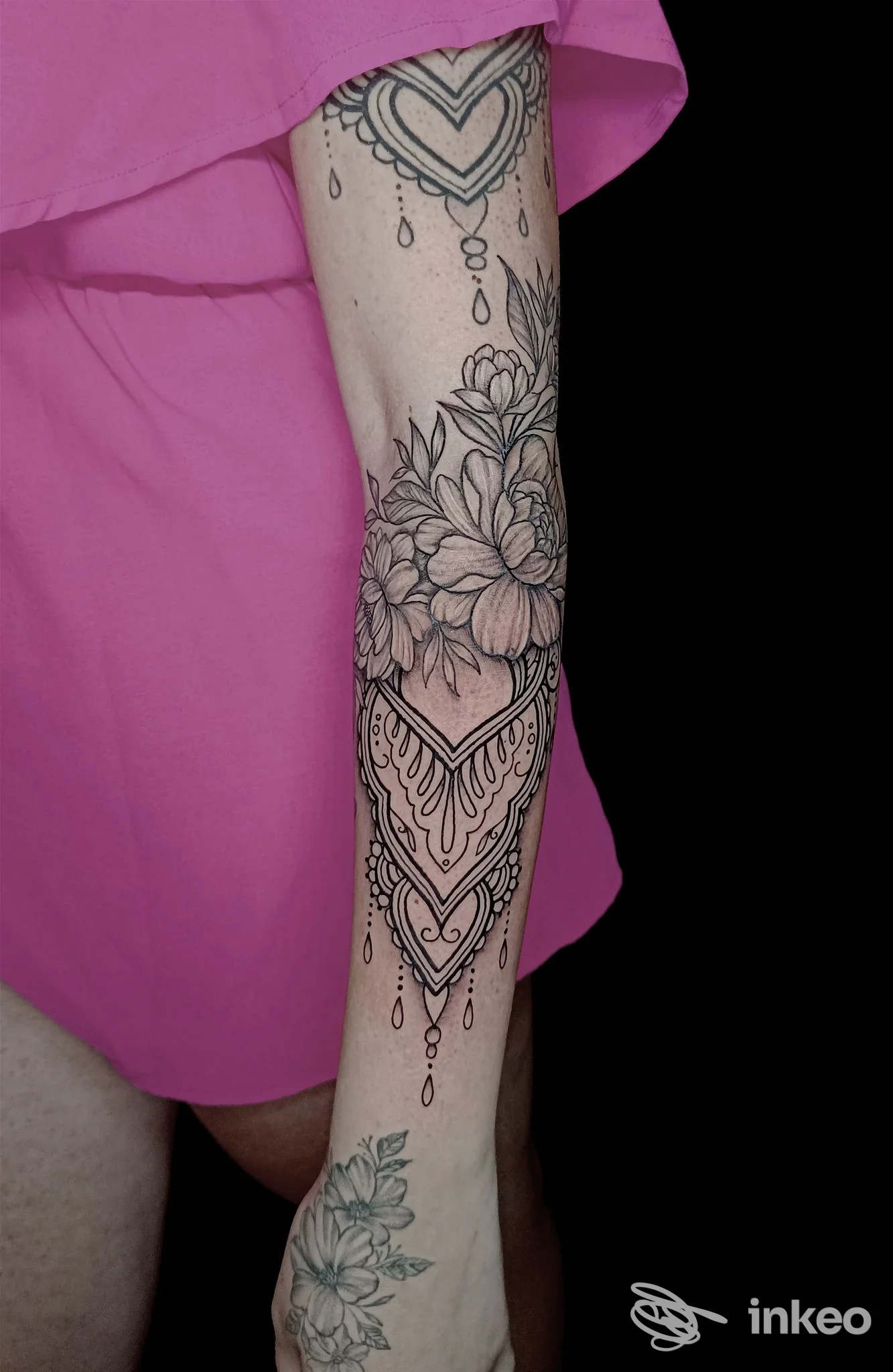 Tattoo su avambraccio sinistro con motivi floreali, catene a perline e cuori, sfumato in stile linework; sfondo nero.