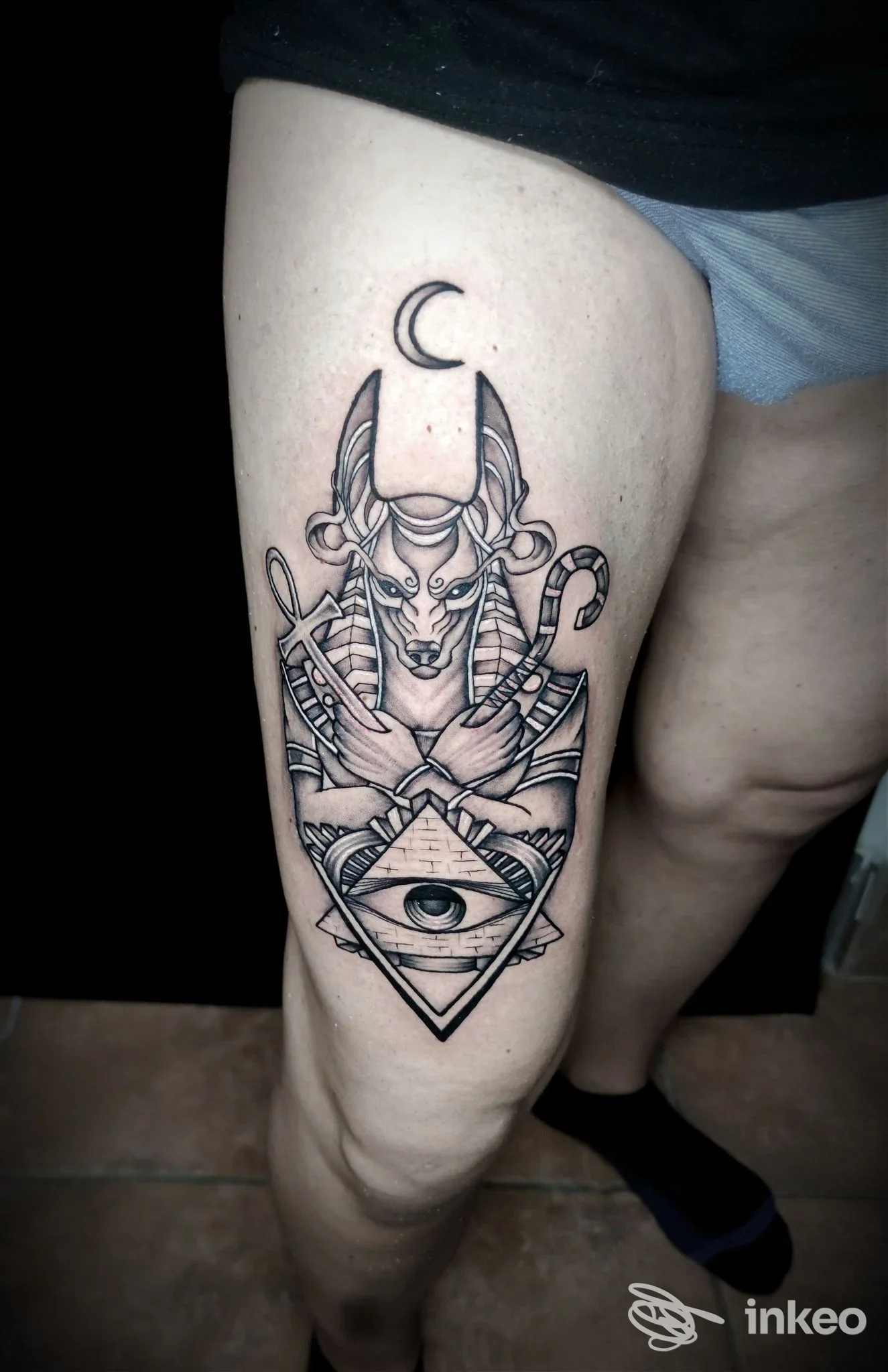 Tattoo visibile sulla coscia, design geometrico con volto demoniaco, triangolo con occhio, luna sopra e simboli incrociati. realizzato direttamente sulla pelle.