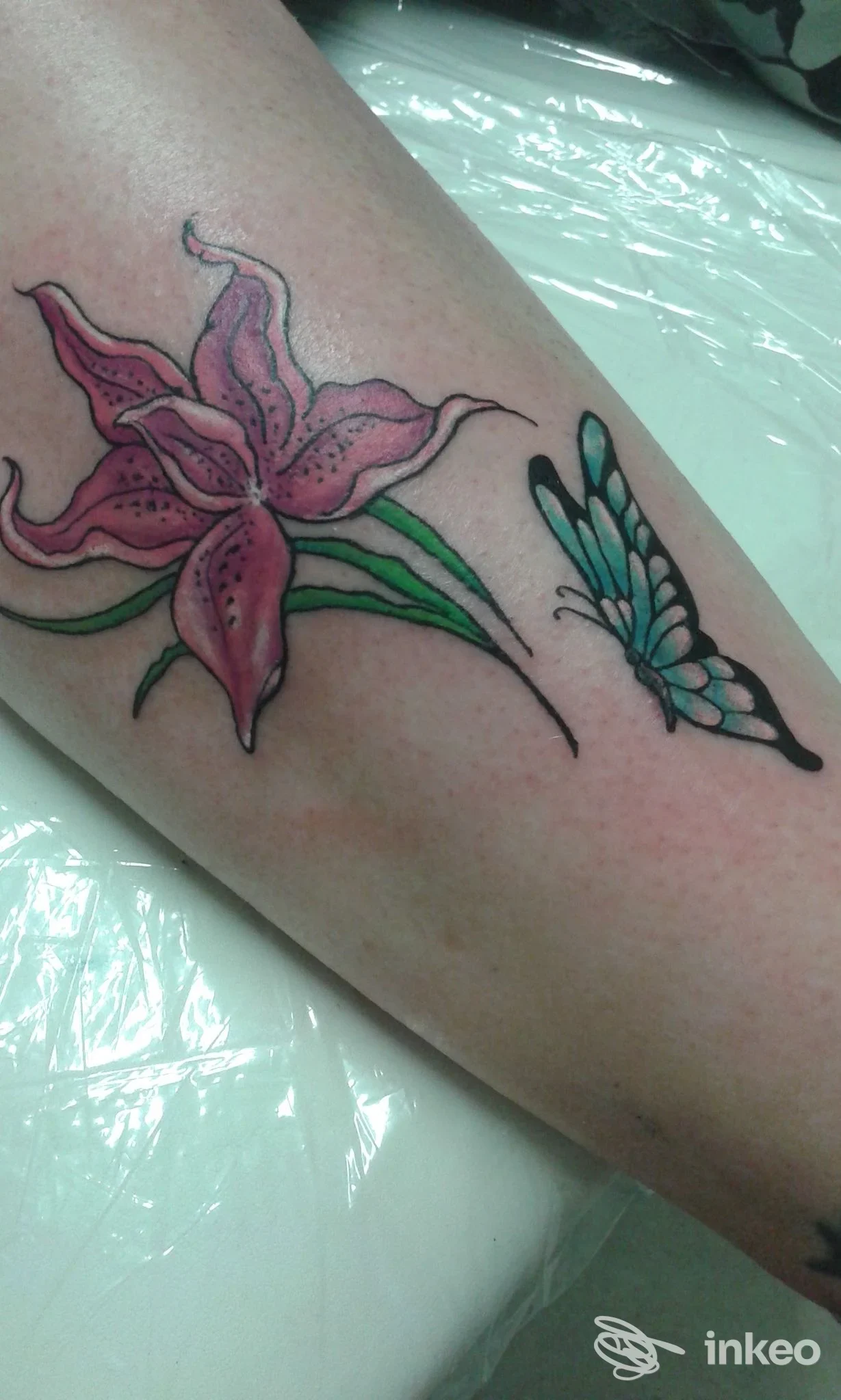 Un tatuaggio su braccio raffigura un fiore rosa e una farfalla azzurra, realizzato sulla pelle. Nessun volto visibile, contesto di studio tatuaggi. Peonia Tattoo