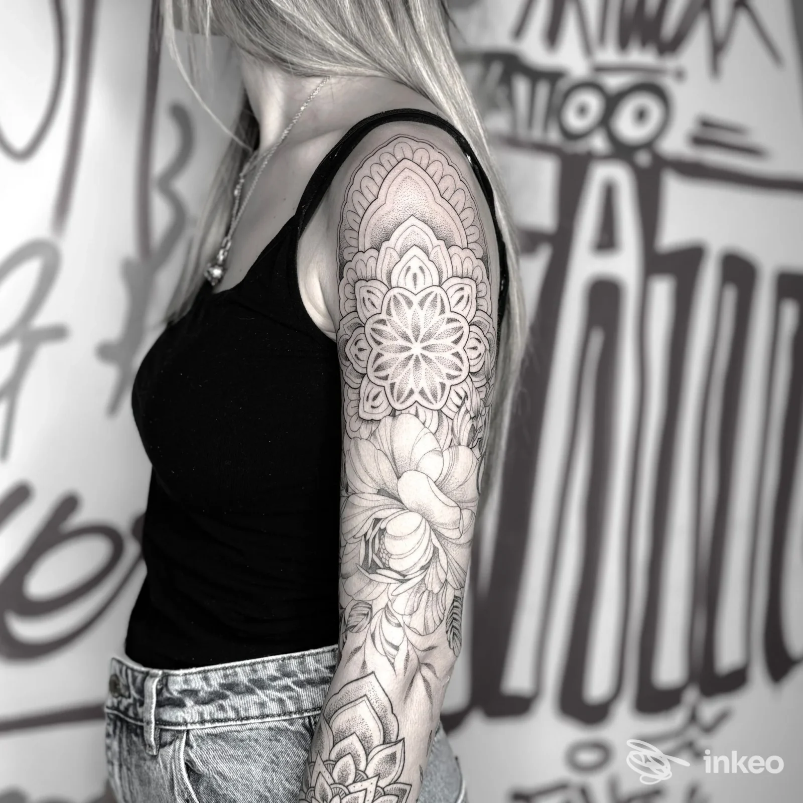 Tattoo sul braccio di una persona, sleeve con motivi floreali e geometrici, in bianco e nero, sfondo con graffiti, ritratto parziale. studio non specificato.