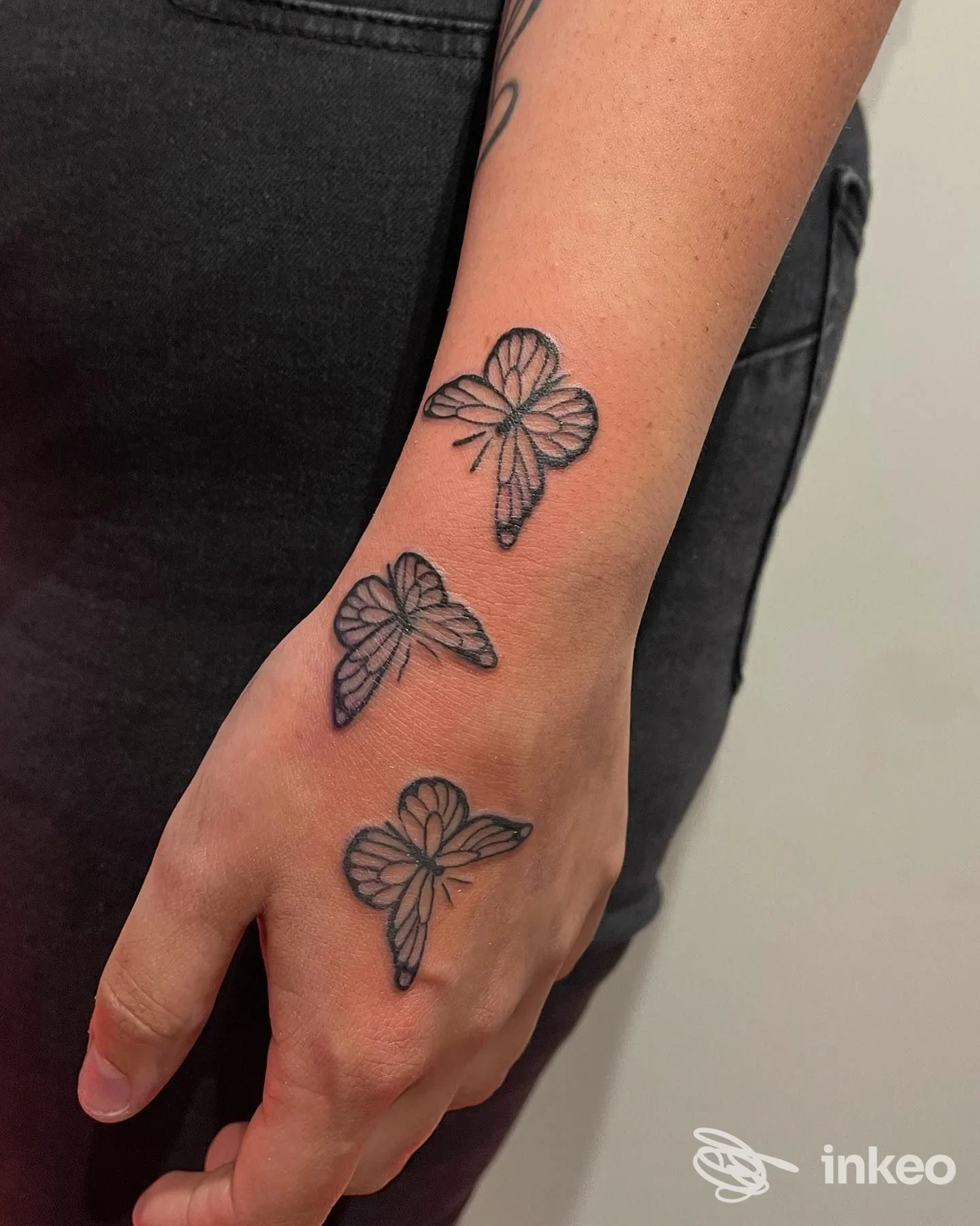 Tre piccoli tatuaggi a forma di farfalla sul dorso della mano, linea sottile nera su pelle chiara, visible vicino al polso.