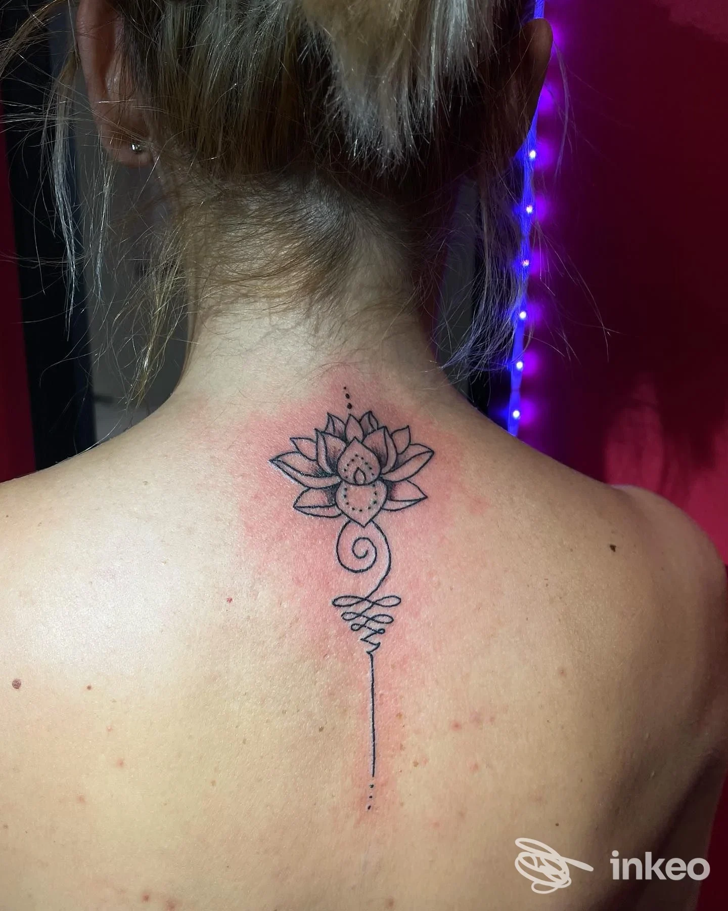 Tattoo sul collo-nuca con loto stilizzato e linea che scende lungo la schiena, eseguito su pelle, colori nero e sfumature.