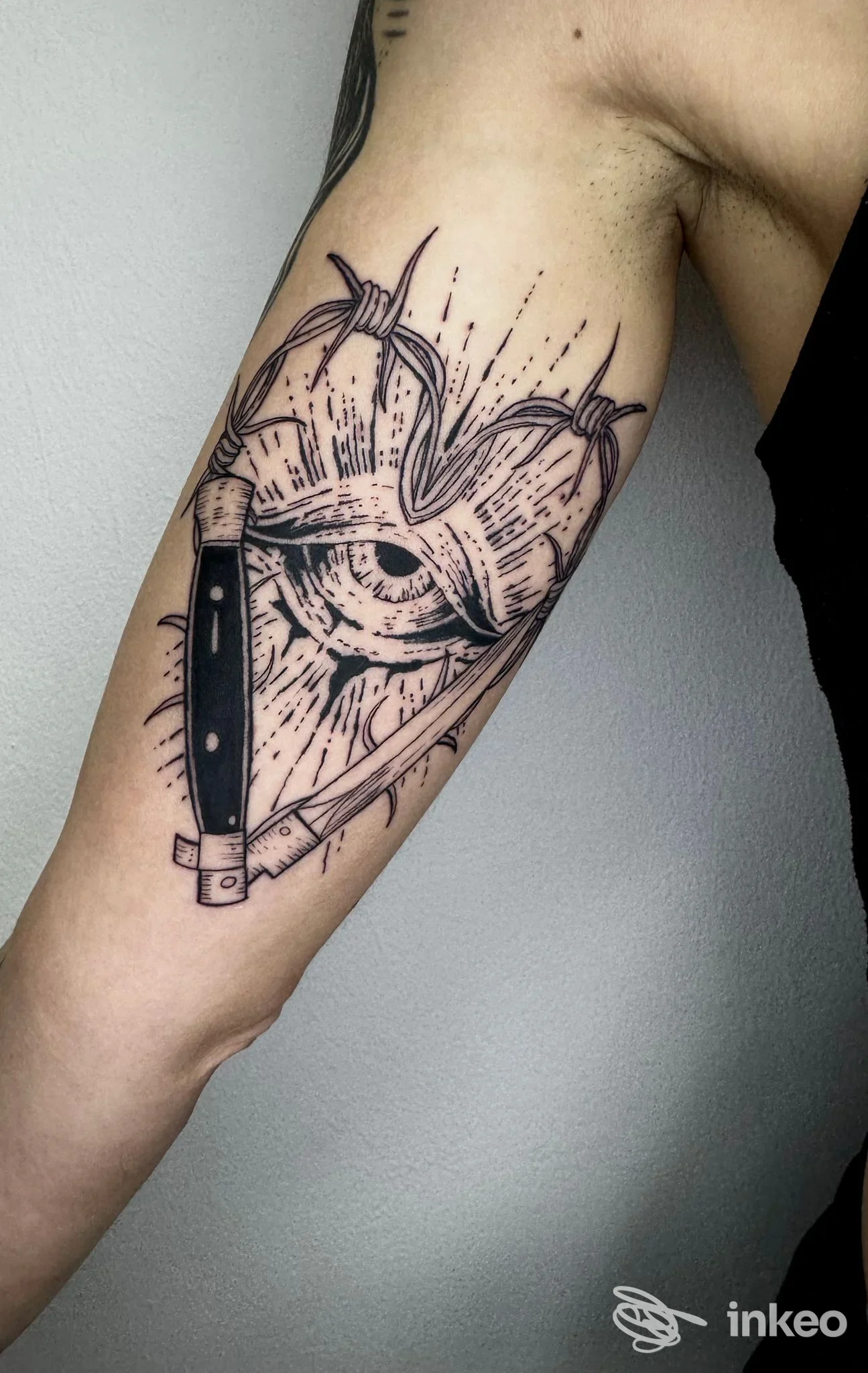 Tattoo visibile sul braccio superiore: disegno a linework con lama e occhio al centro, bordi netti e ombreggiature minime.
