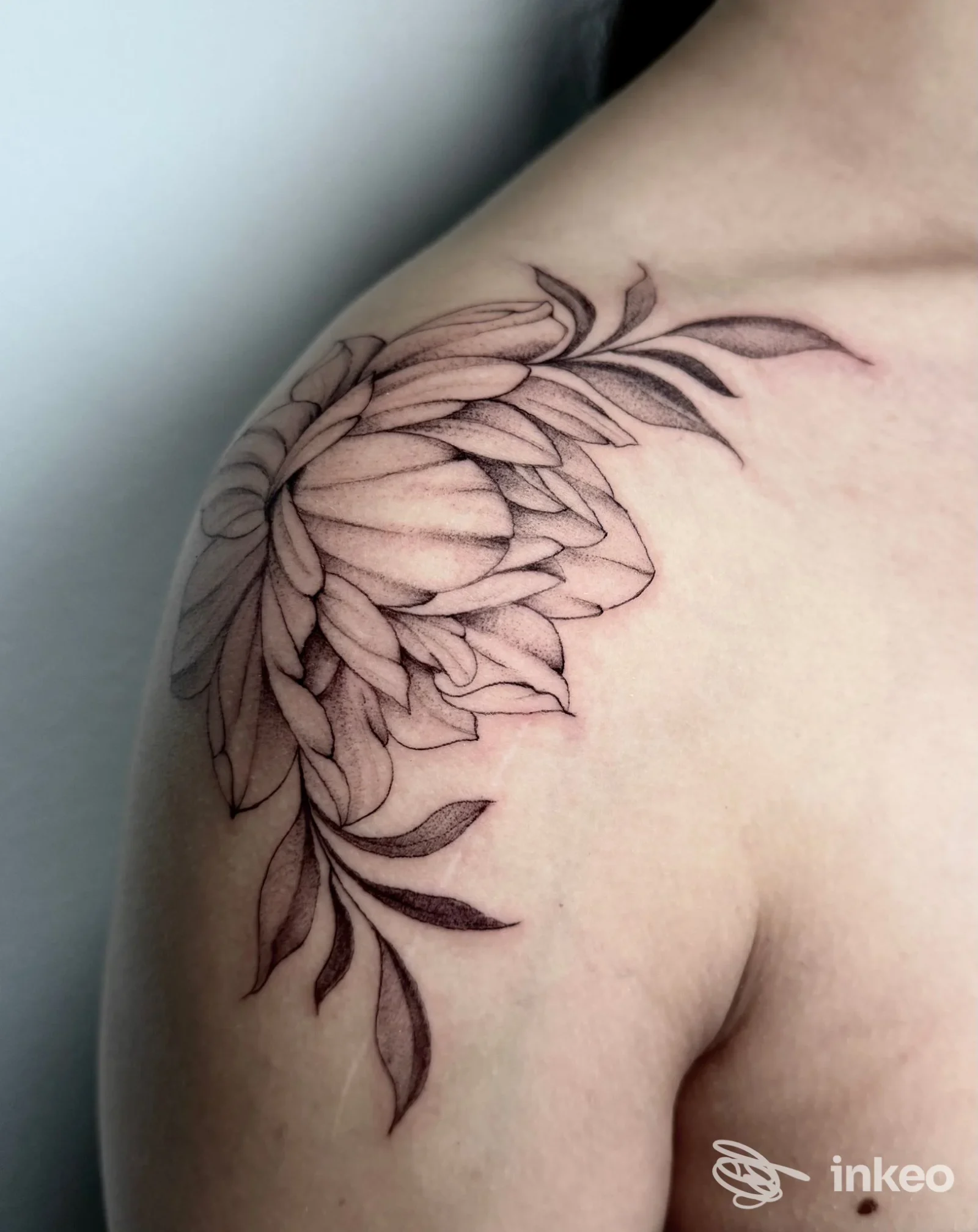 Tattoo floreale stilizzato sulla spalla sinistra, disegno a linee fini e shading delicato, visibile sulla pelle di una persona; tatuaggio reale senza elementi di interfaccia.