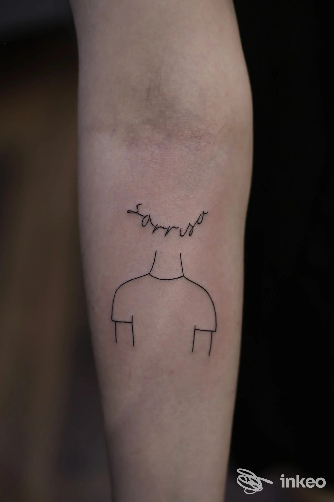 Tattoo minimalista su avambraccio: linea nera che suggerisce una figura con spalle scoperte, senza dettagli del viso. lo sfondo è scuro, si legge la firma o parola sopra la testa. Studio Mr Mike, Canzo (CO).