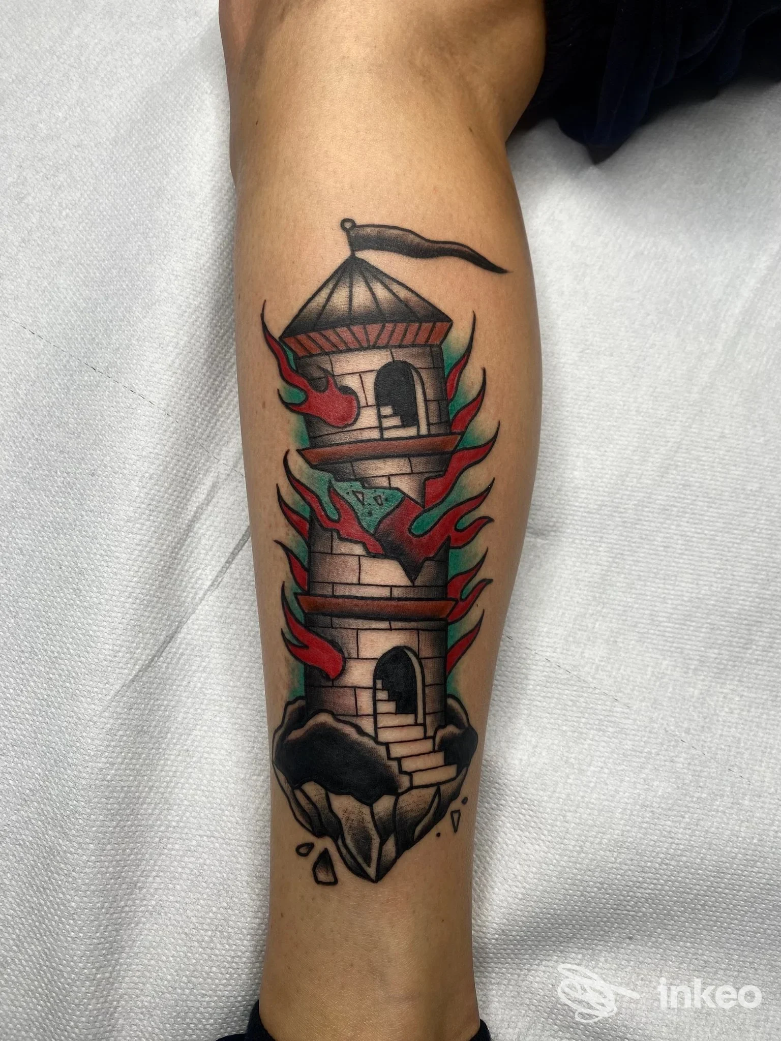 Tattoo a torre a più piani avvolta da fiamme rosse e verdi sull'avambraccio sinistro, con dettagli in nero e riempimenti colorati, scatto singolo.