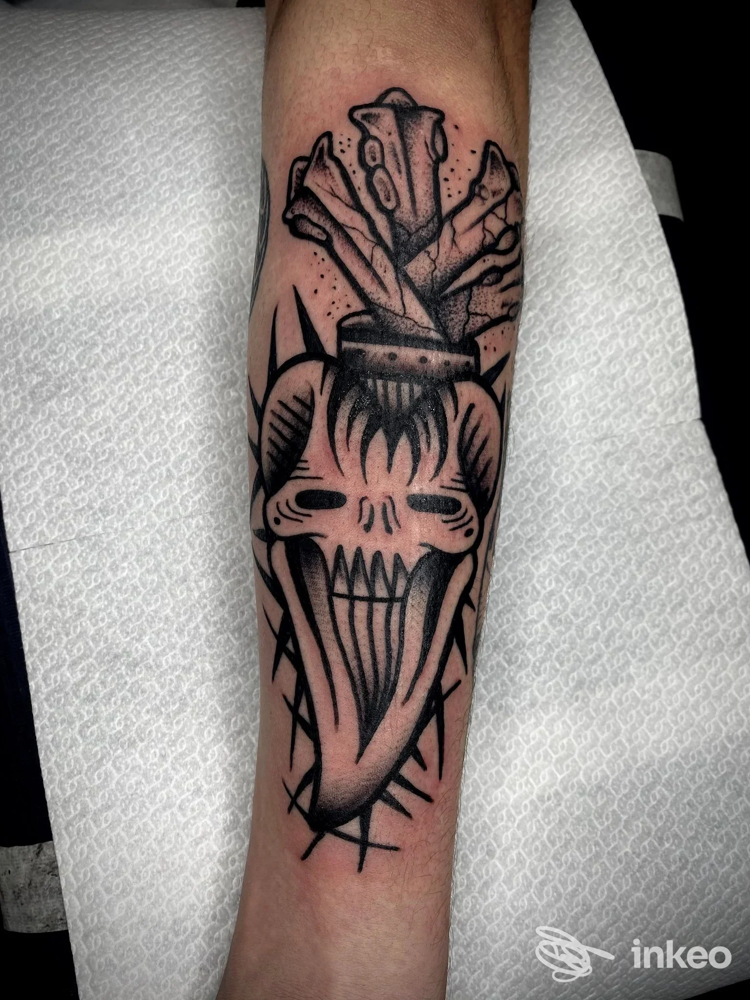 Tatuaggio su avambraccio raffigurante una testa scheletrica stilizzata con linguaccia, contorni neri marcati e motivi a frecce attorno, realizzato su pelle; studio Broken Bones Tattoo.