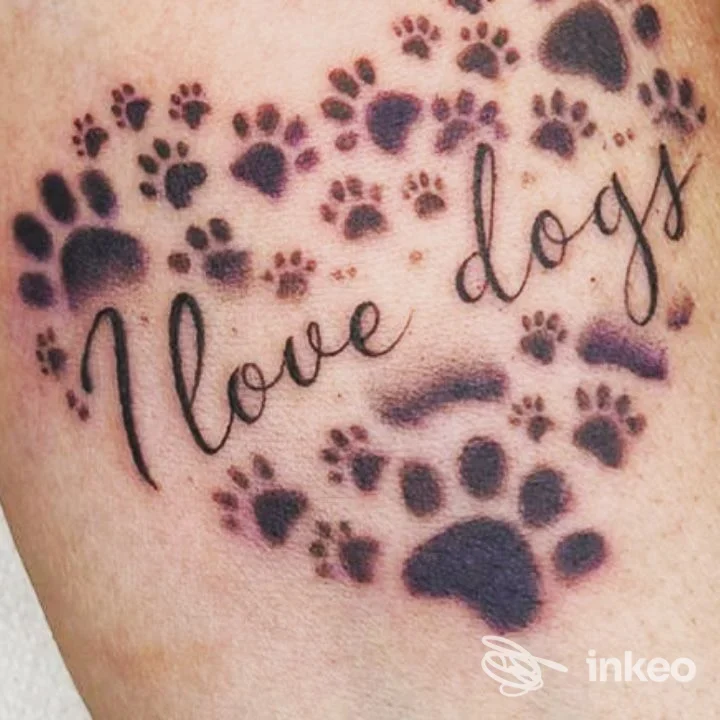 Tatuaggio su pelle con numerose impronte di zampa nere attorno a una scritta corsiva che dice 'I love dogs'.