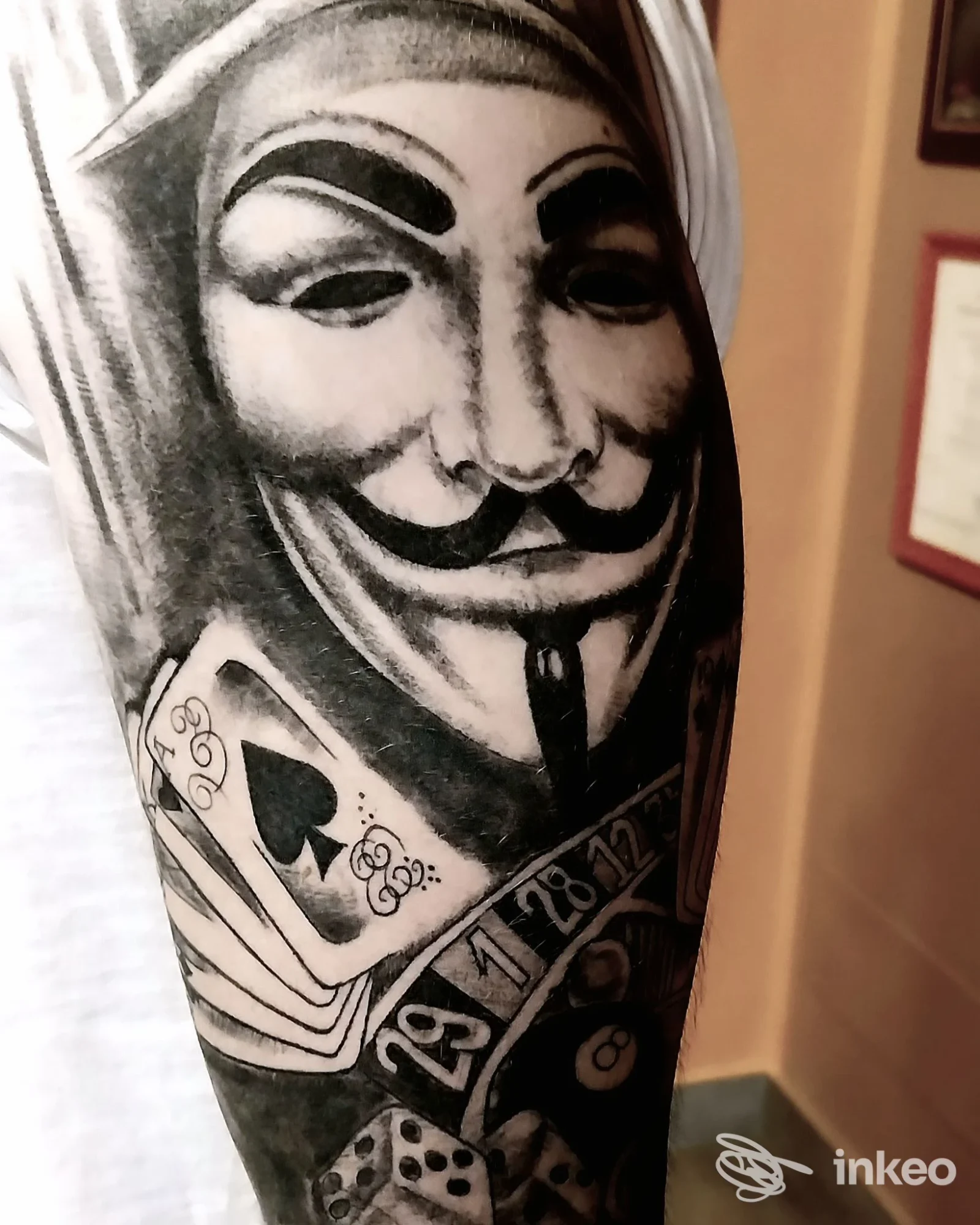 Tattoo in bianco e nero sull'avambraccio: una maschera tipo Guy Fawkes con carte da gioco e dadi, visibile una porzione di pelle; eseguito in studio, nessun overlay visibile.