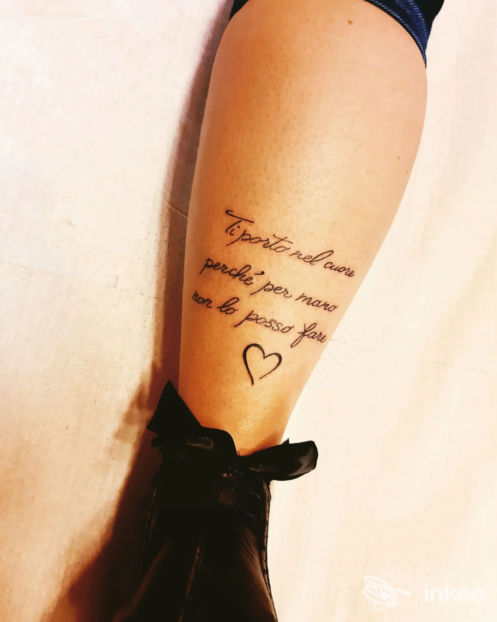 Una gamba nuda con un tatuaggio di testo in corsivo e un piccolo cuore disegnato sulla parte anteriore del polpaccio, con una scarpa nera in primo piano.