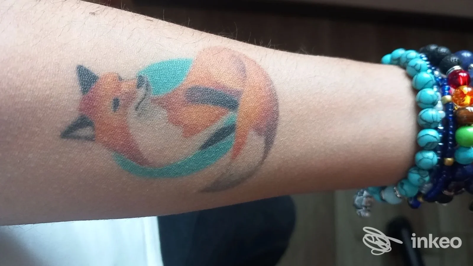 Tatuaggio colorato di una volpe stilizzata sul polso, con braccialetti colorati al polso della persona.