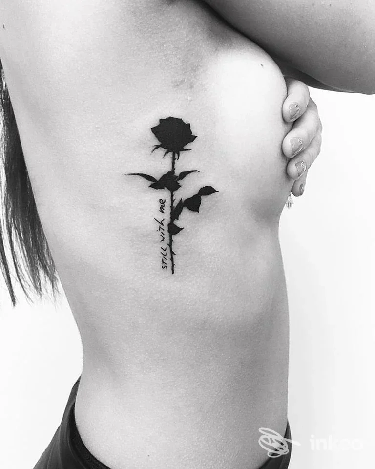 Tattoo realistico di una rosa nera sul fianco sinistro, lungo la gabbia toracica, con testo verticale sul gambo; visibile solo il fianco della persona.