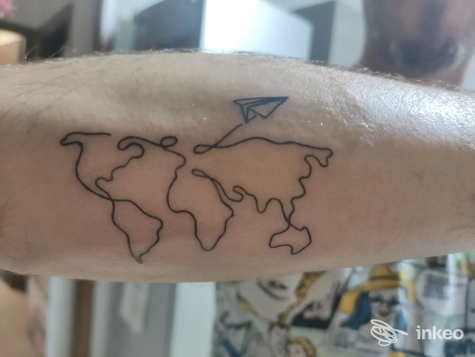 Avambraccio con tatuaggio stilizzato raffigurante una mappa mondiale e un aereo di carta sospeso sopra di essa, linee nere su pelle chiara.