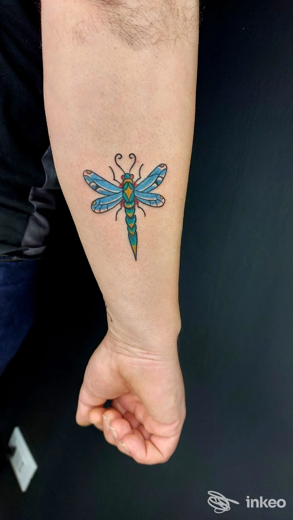 Avambraccio sinistro con tatuaggio colorato di una libellula, ali blu e corpo verde-giallo, in primo piano.
