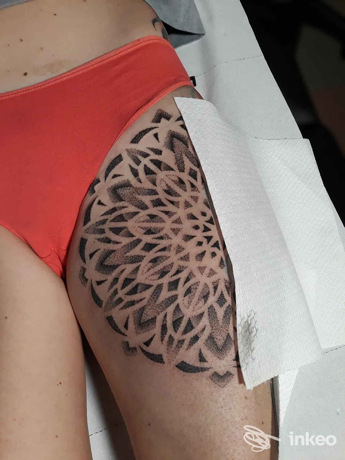 Tattoo geometrico a motivo floreale sulla coscia sinistra di una persona, con dettagli neri netti e sfondo non visibile.
