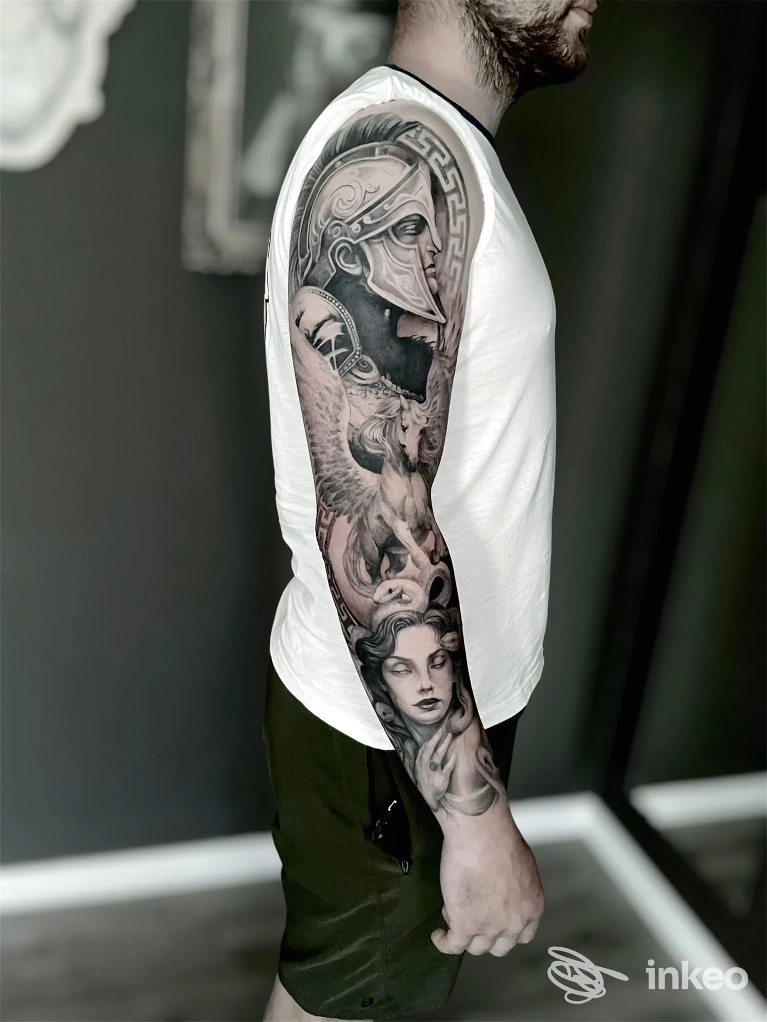Veduta laterale di una persona con una manica tatuata sul braccio sinistro, tatuaggi in bianco e nero con elmo, figure decorative e volti femminili, indossa una maglietta bianca e sfondo scuro.