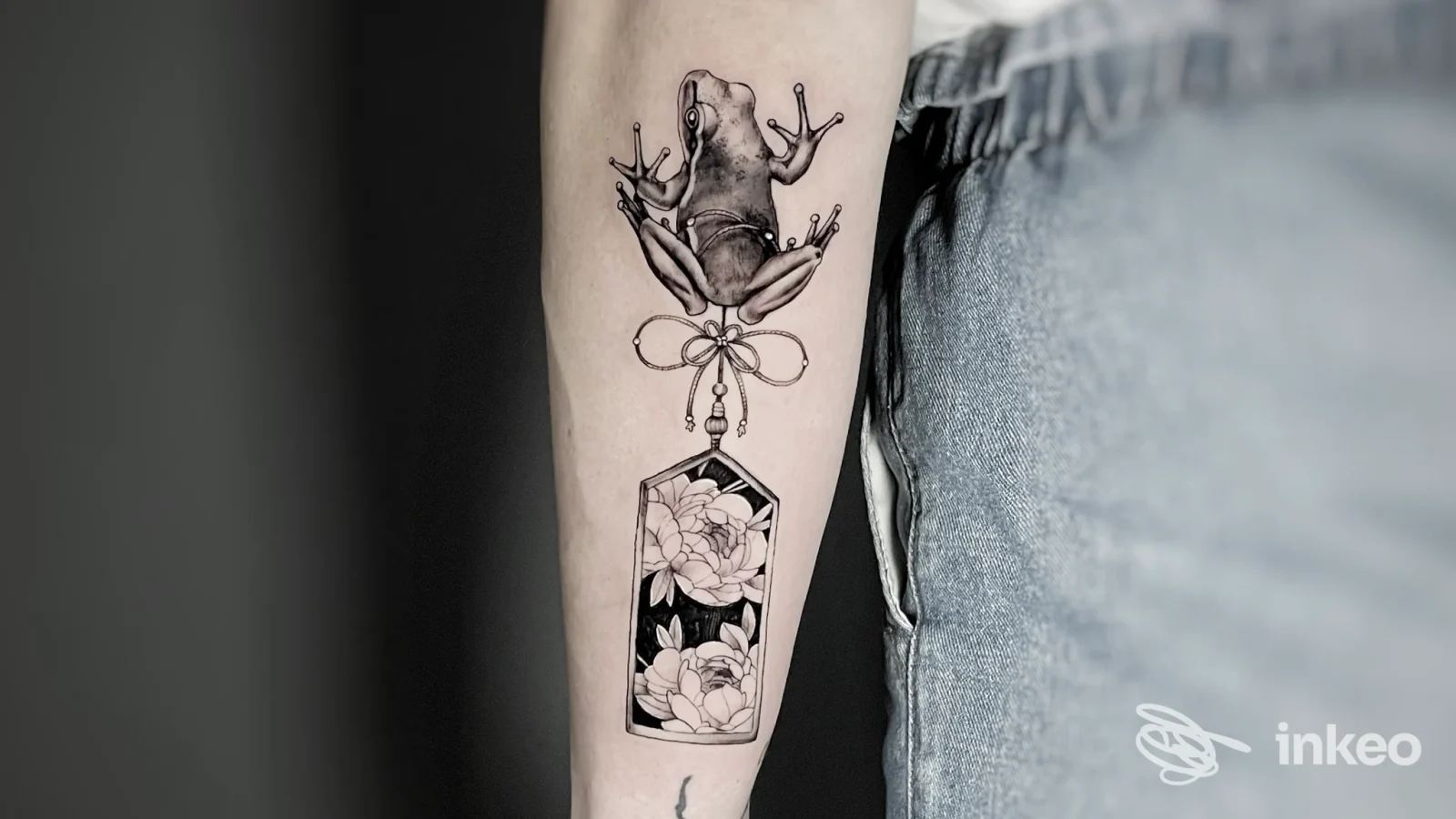 Tattoo visibile sull'avambraccio: rana con ciondolo e motivo floreale all'interno di una placca rettangolare, eseguito sulla pelle.