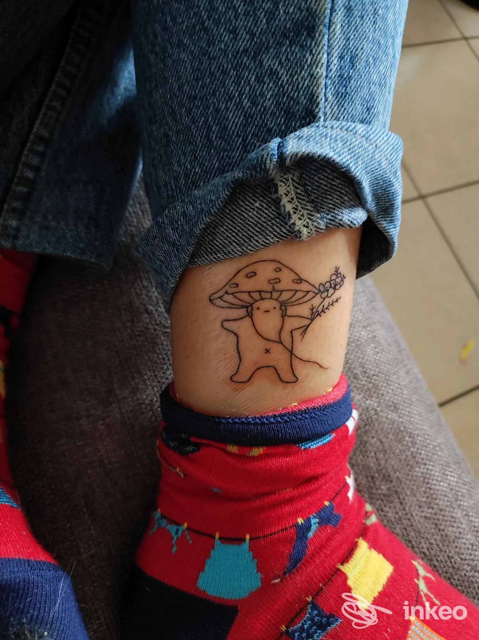 Tatuaggio realizzato a linea nera su pelle della caviglia, raffigurante una piccola creatura stilizzata con cappello, a fianco un fiore; jeans arricciati sullo sfondo.
