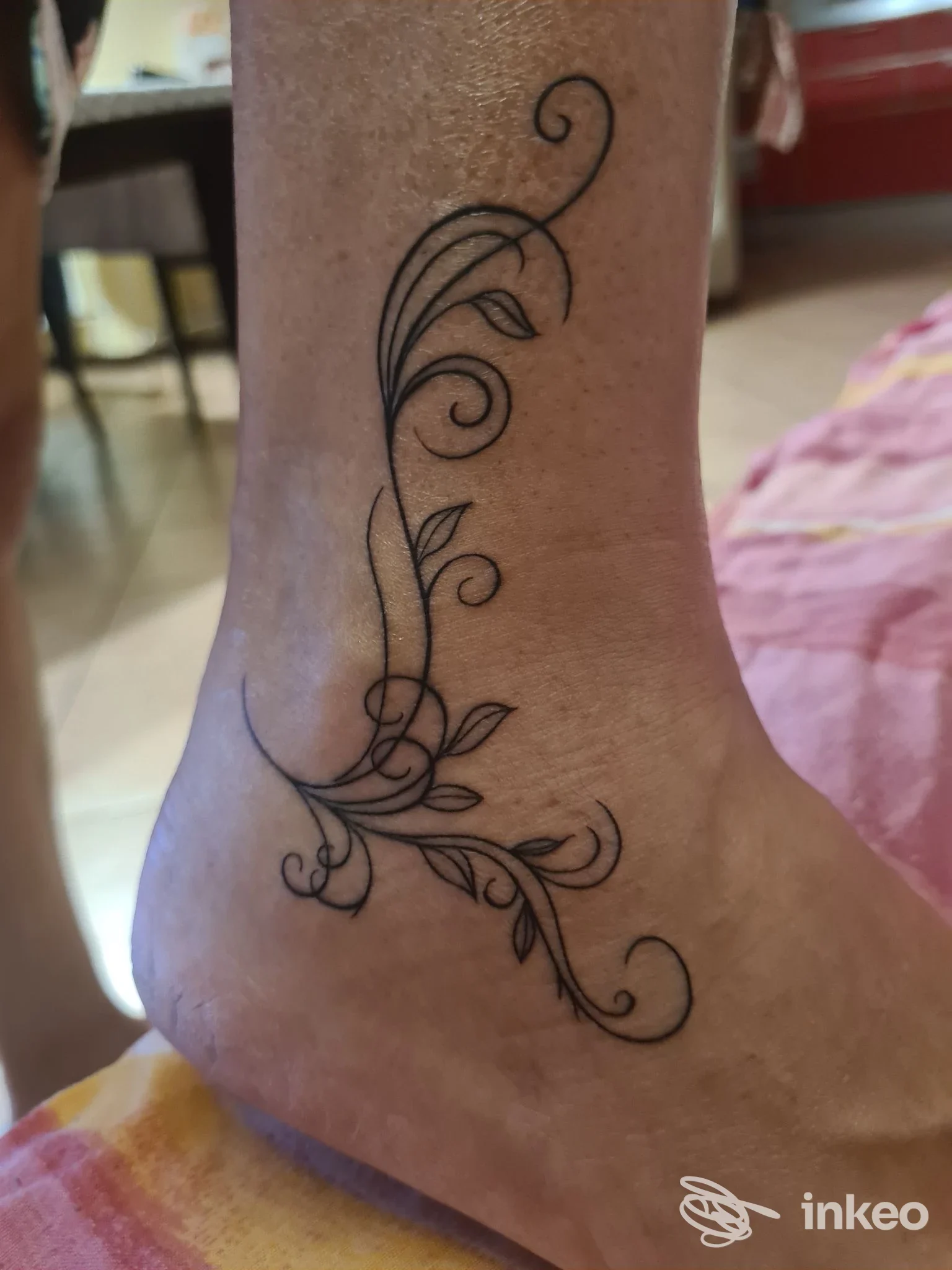 Tatuaggio florale stilizzato lungo la caviglia su pelle reale, visibile solo la parte esterna della gamba e piede, senza volto inquadrato.