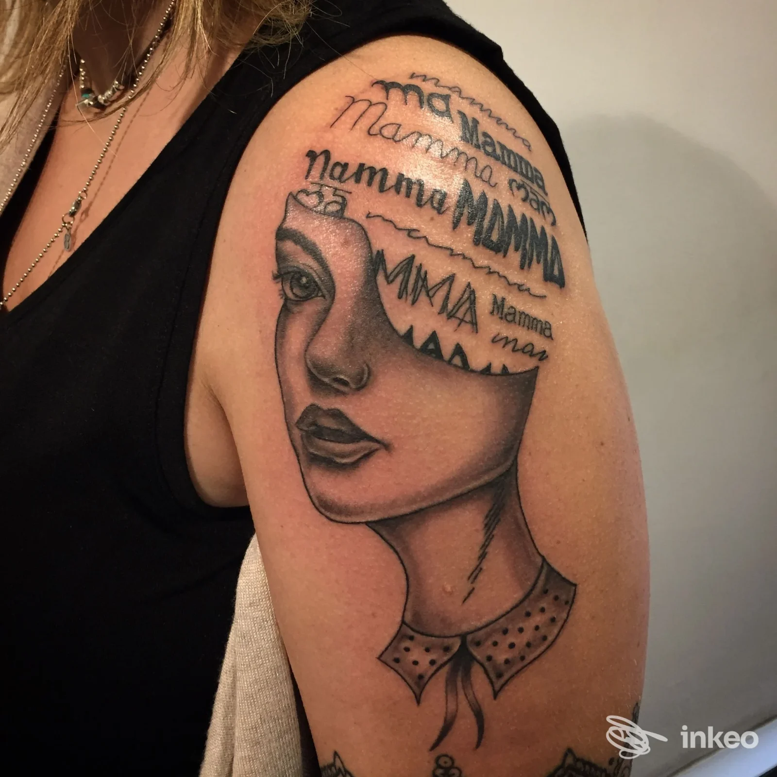 Tattoo sulla spalla raffigurante un volto femminile stilizzato con il testo ricorrente Mamma, visibile su pelle reale senza watermark o overlay sociale.