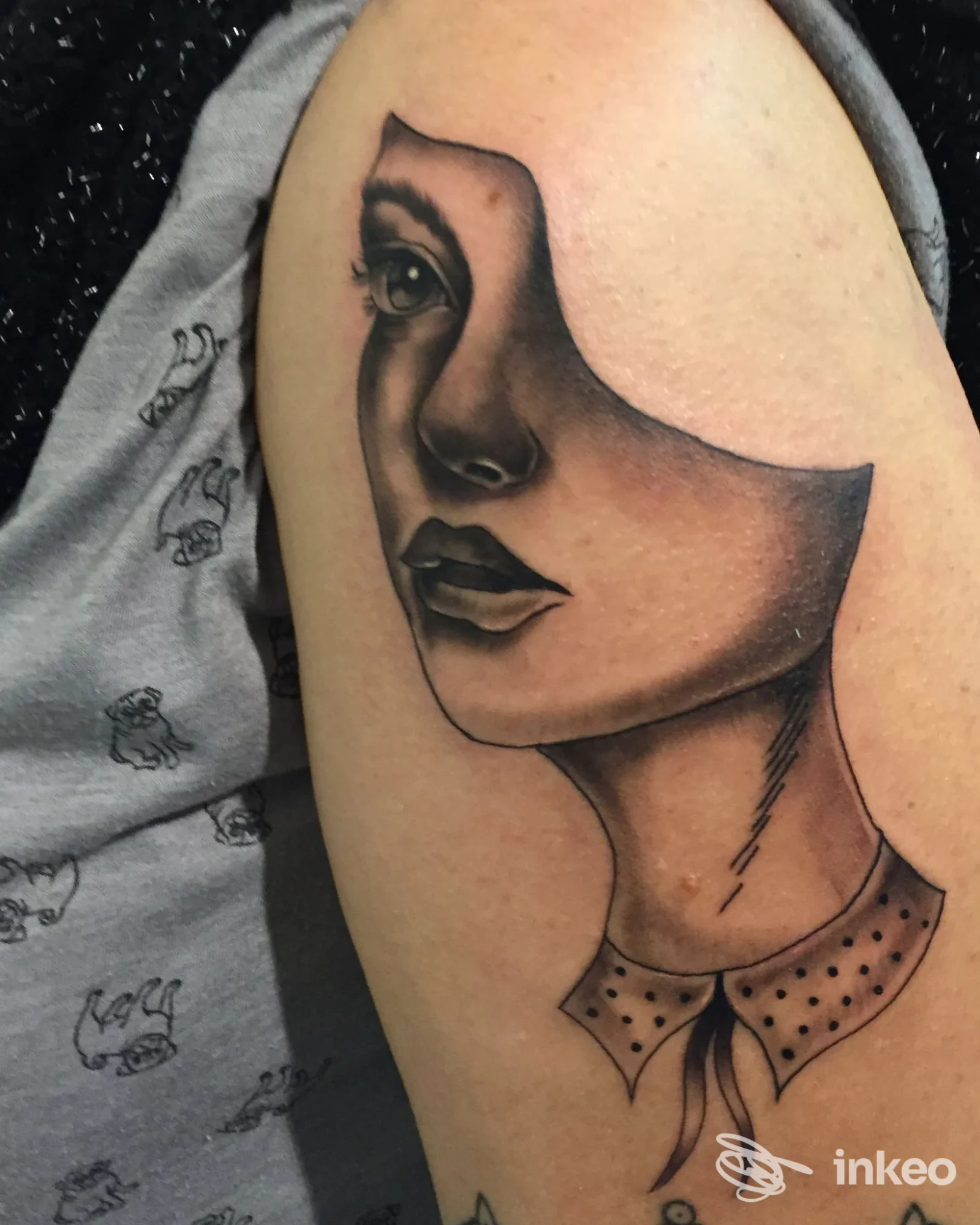 Tattoo sull'avambraccio raffigurante un volto femminile stilizzato, contorni netti e sfumature, con maglia fantasia sul colletto.